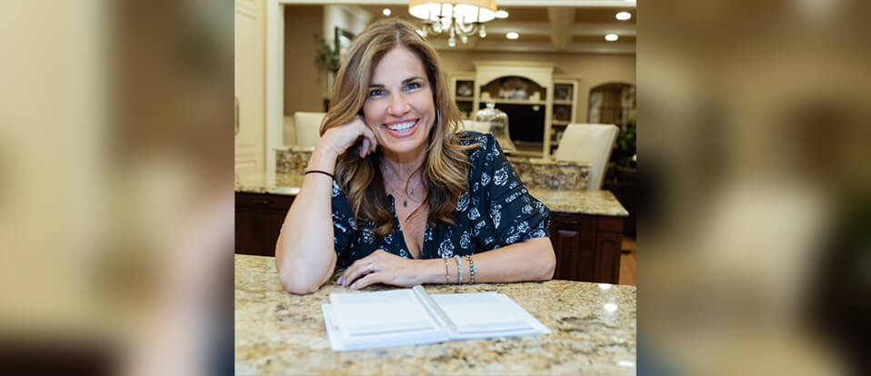 Julie Traina smiles with her head in her hand while sitting at a kitchen table with a notebook.