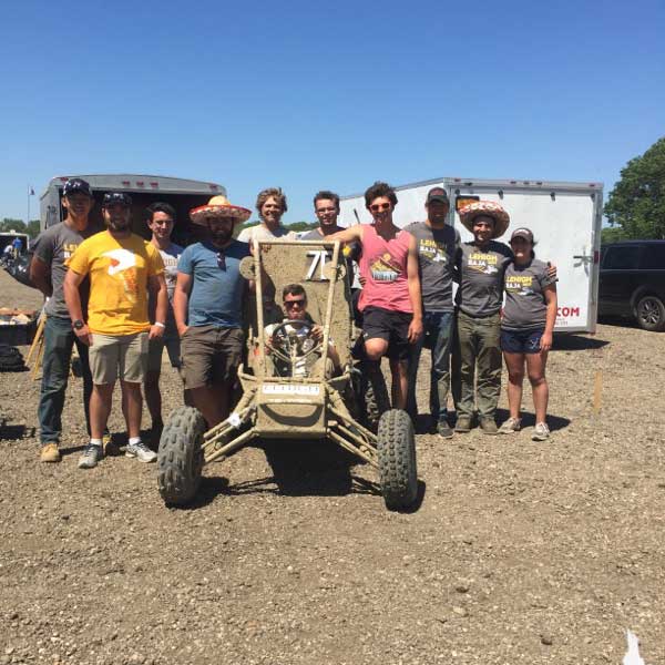 A group of people gather around a beige, two-wheeled vehicle. Justin sits behind the wheel of the car and smiles.