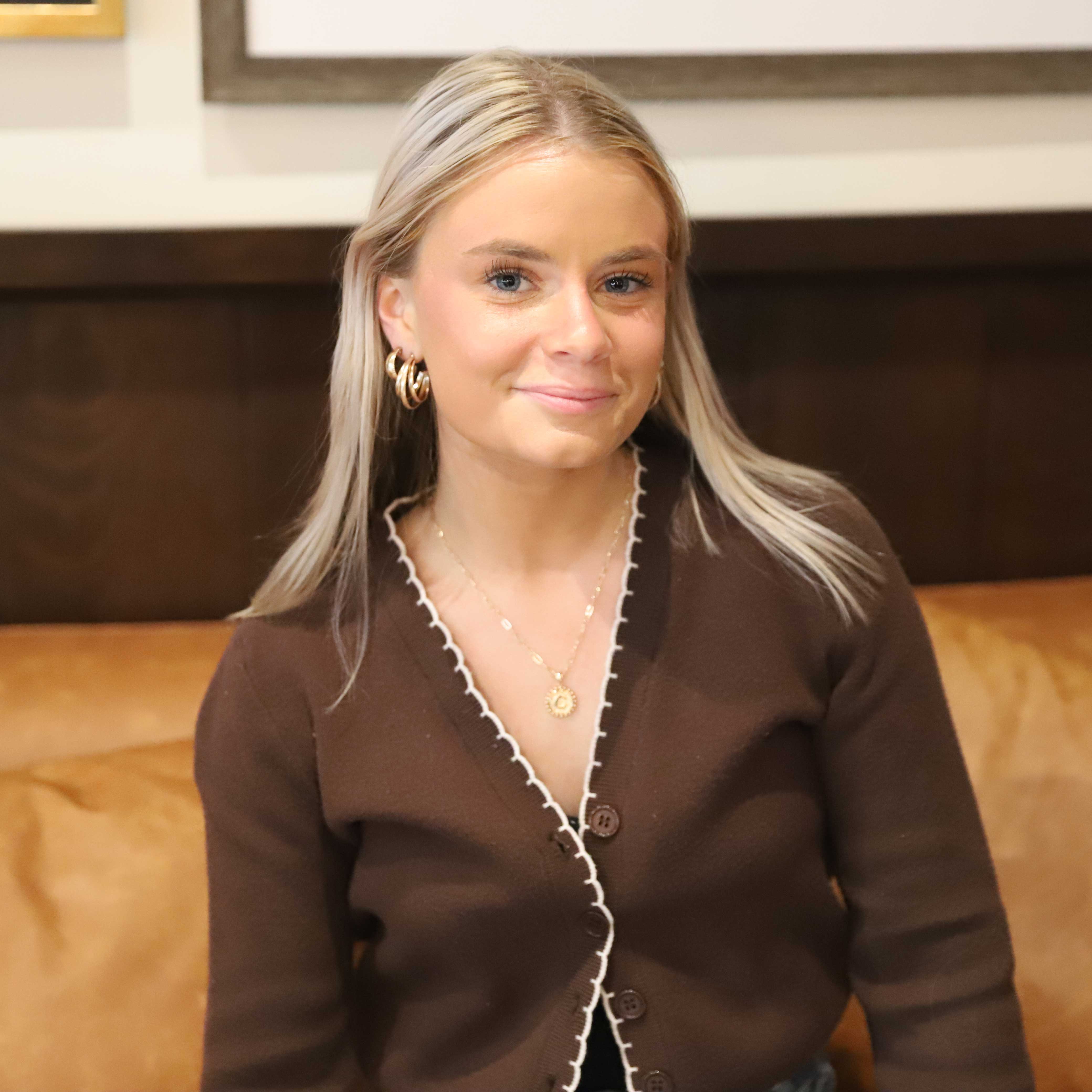 Caitlyn sits on a brown leather couch. She wears a dark brown cardigan; photographs of students are behind her.