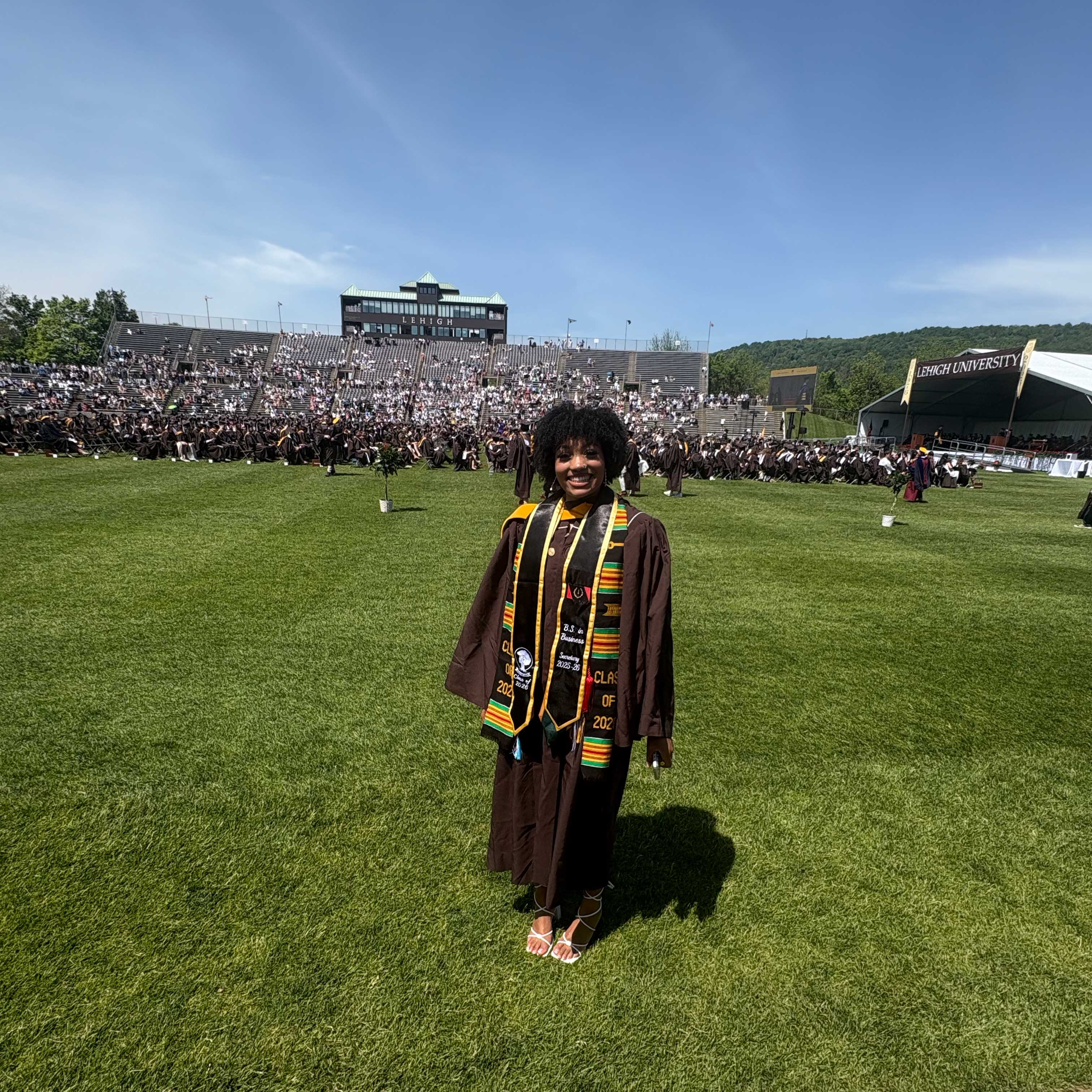 Kamara wears her graduation gown and smiles while standing on the field; behind her, students are visible.
