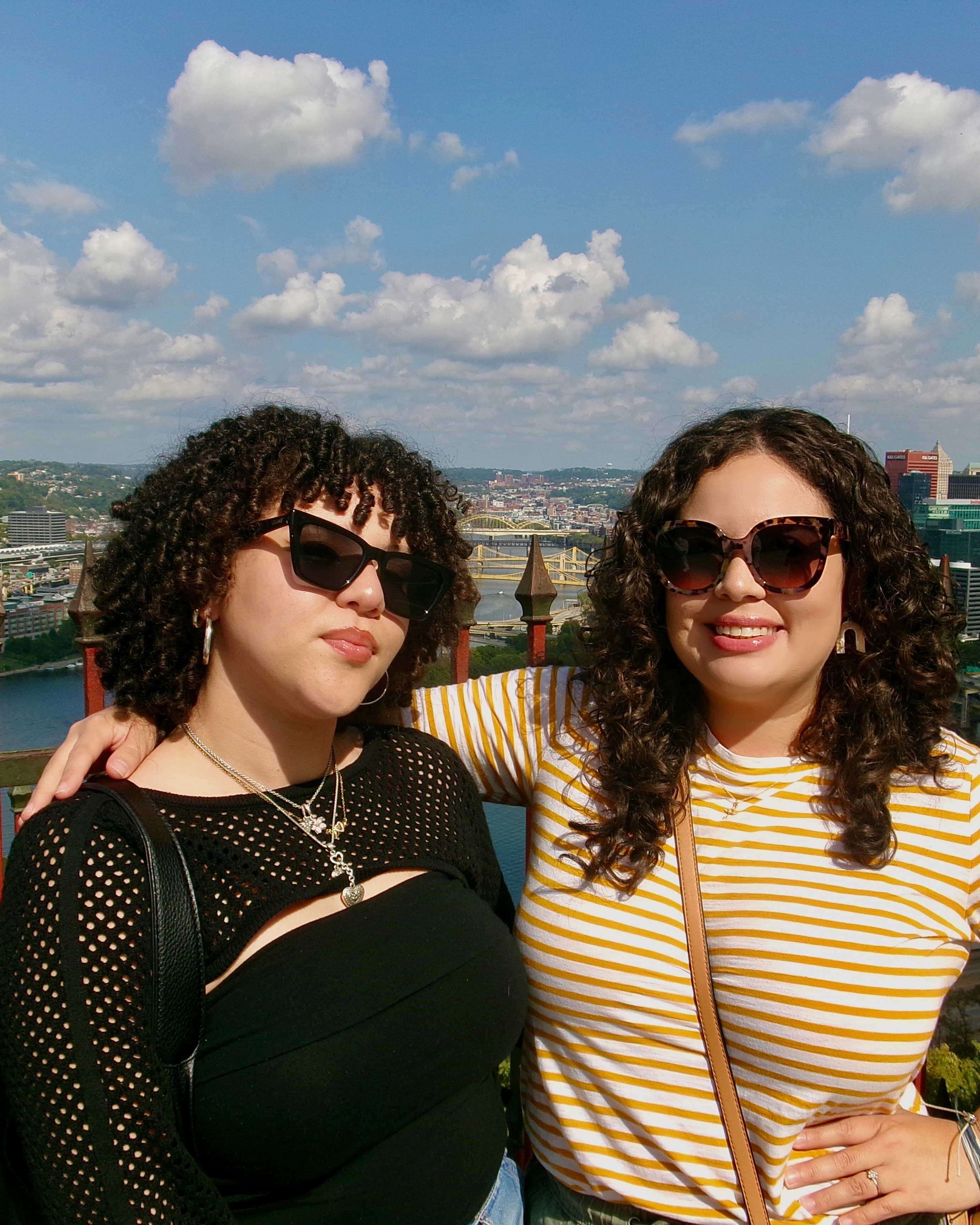 Karina wraps an arm around Kayla's shoulder. They both wear sunglasses. Behind them is a bustling city. The sky is clear.