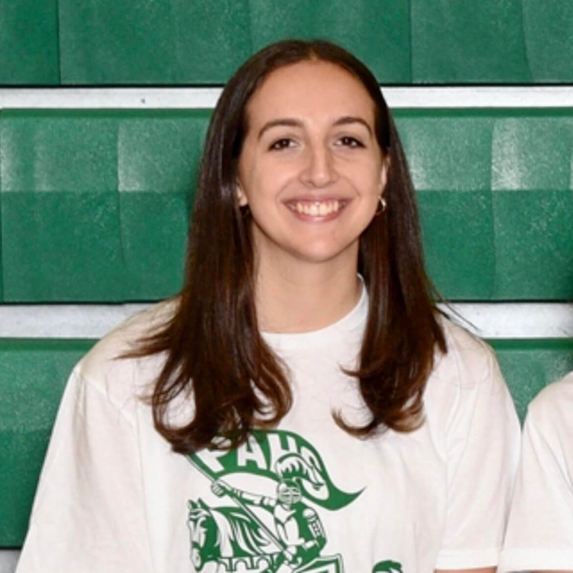Woman smiles for a photo wearing a white t-shirt with a green graphic of a knight on a horse carrying a flag that says "PAHS." 