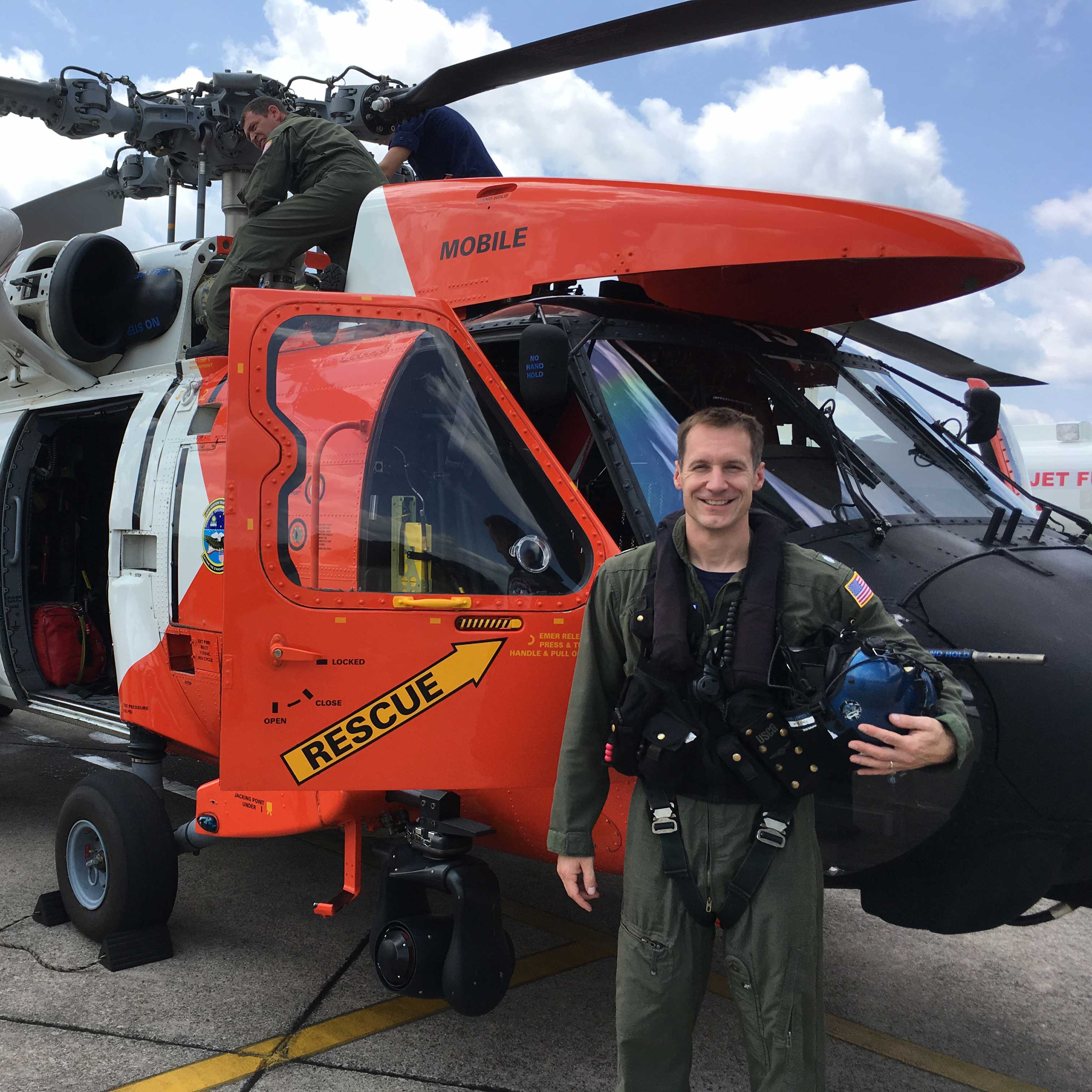 Keith wears a green flight suit and holds his helmet while standing in front of an orange search-and-rescue helicopter. 