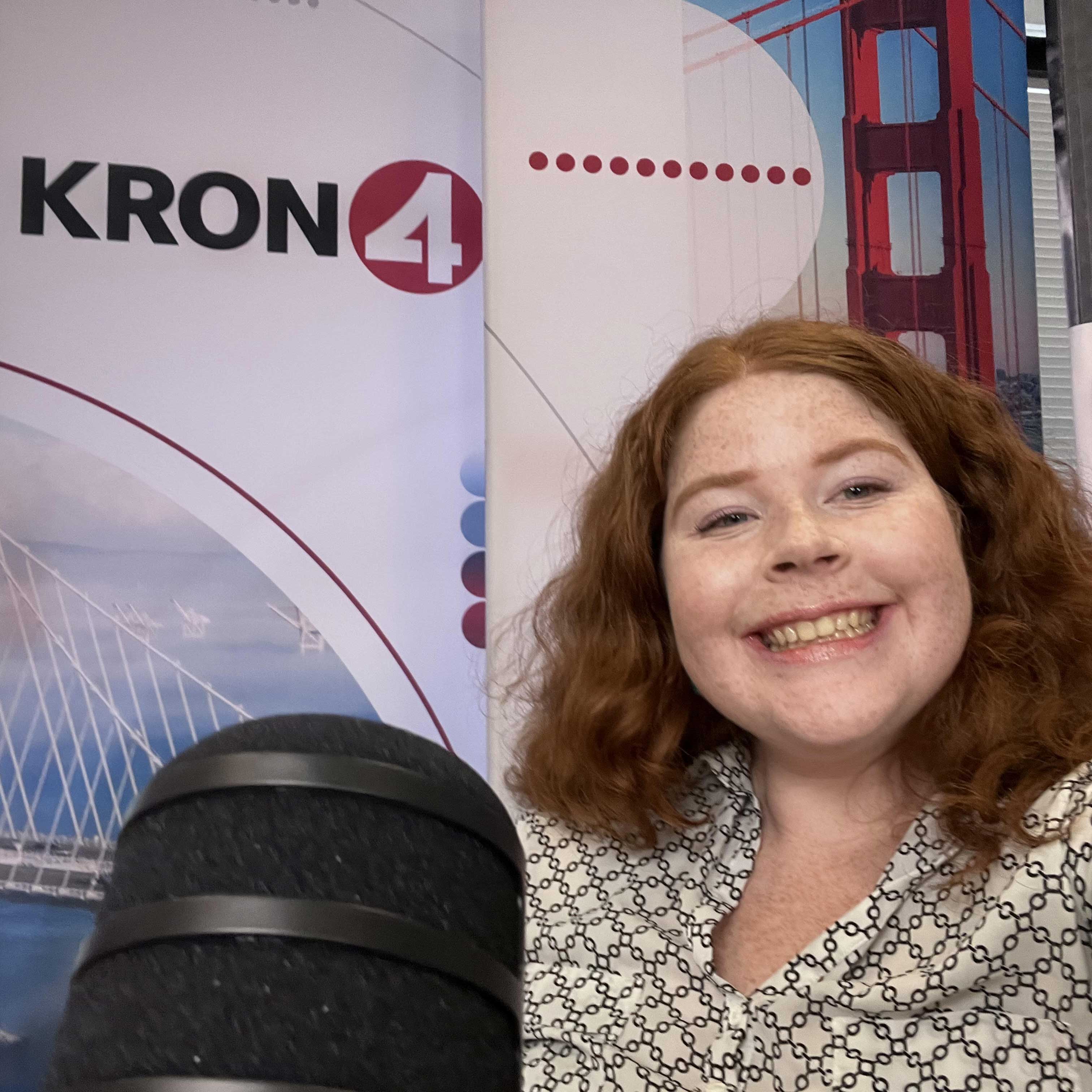 Kendall sits with a black microphone posed in front of them. Behind them is KRON4 news branded background along with city art.