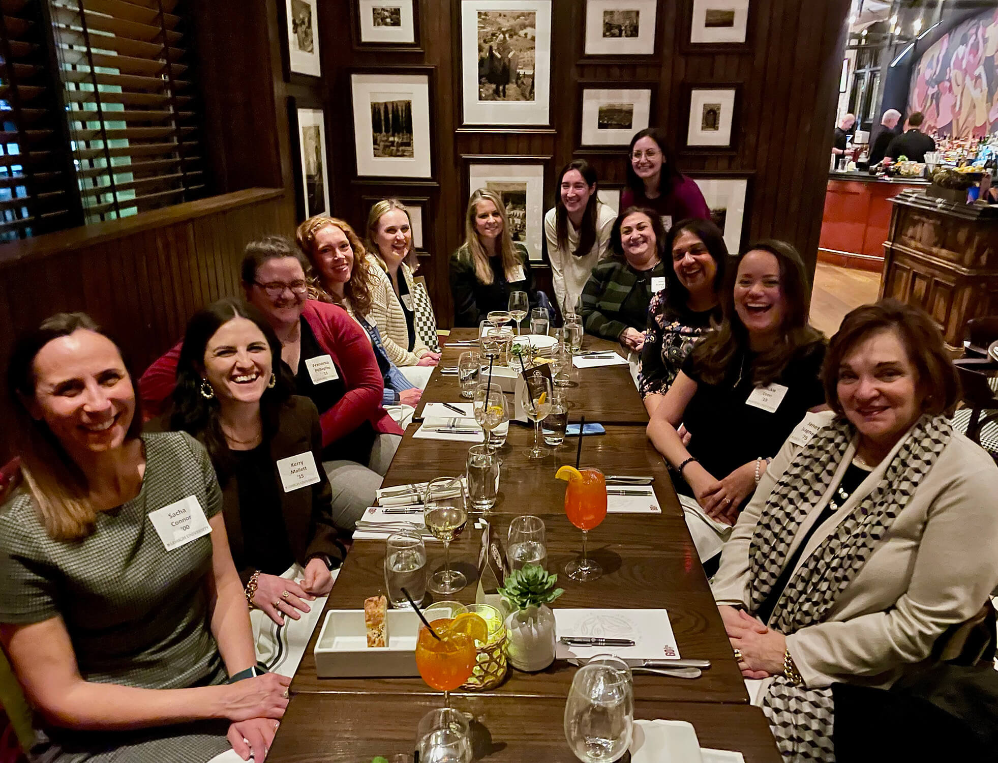 A group of women sit at a long table in a restaurant posing for a photo while 2 at the end squat to join the picture.