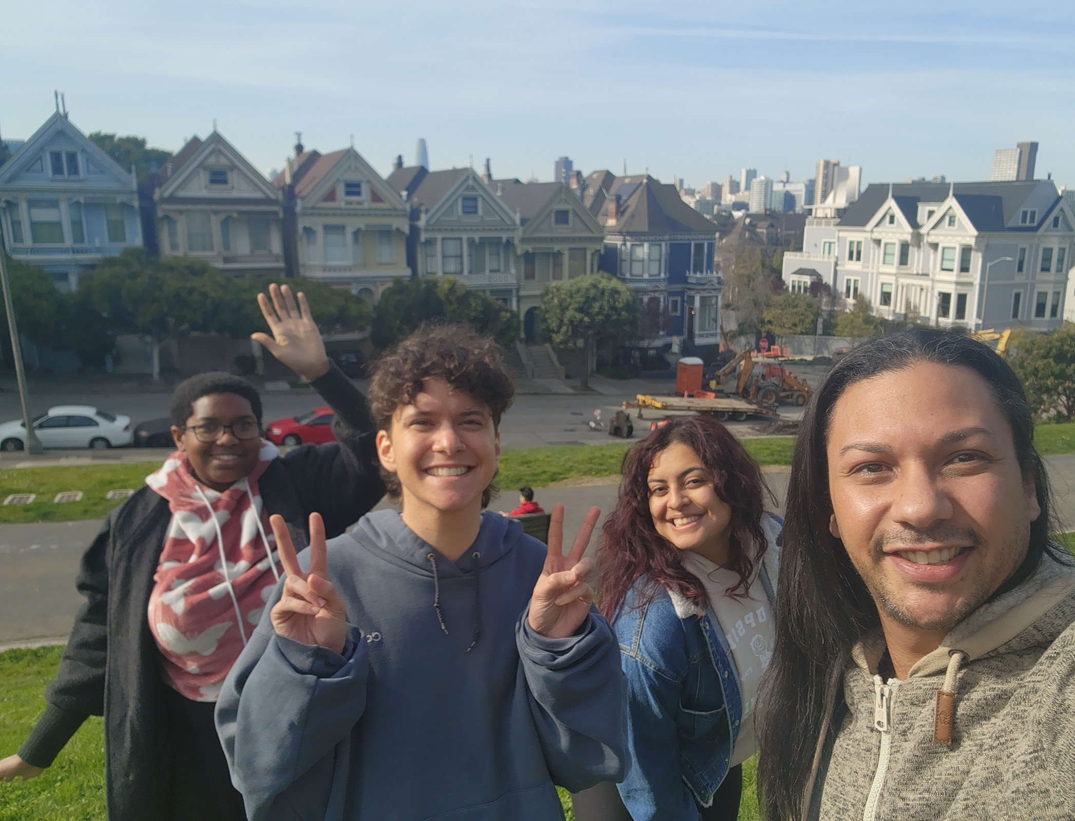 Four people stand in front of iconic San Francisco street with Victorian-style houses, smiling while one makes peace signs with his hands..