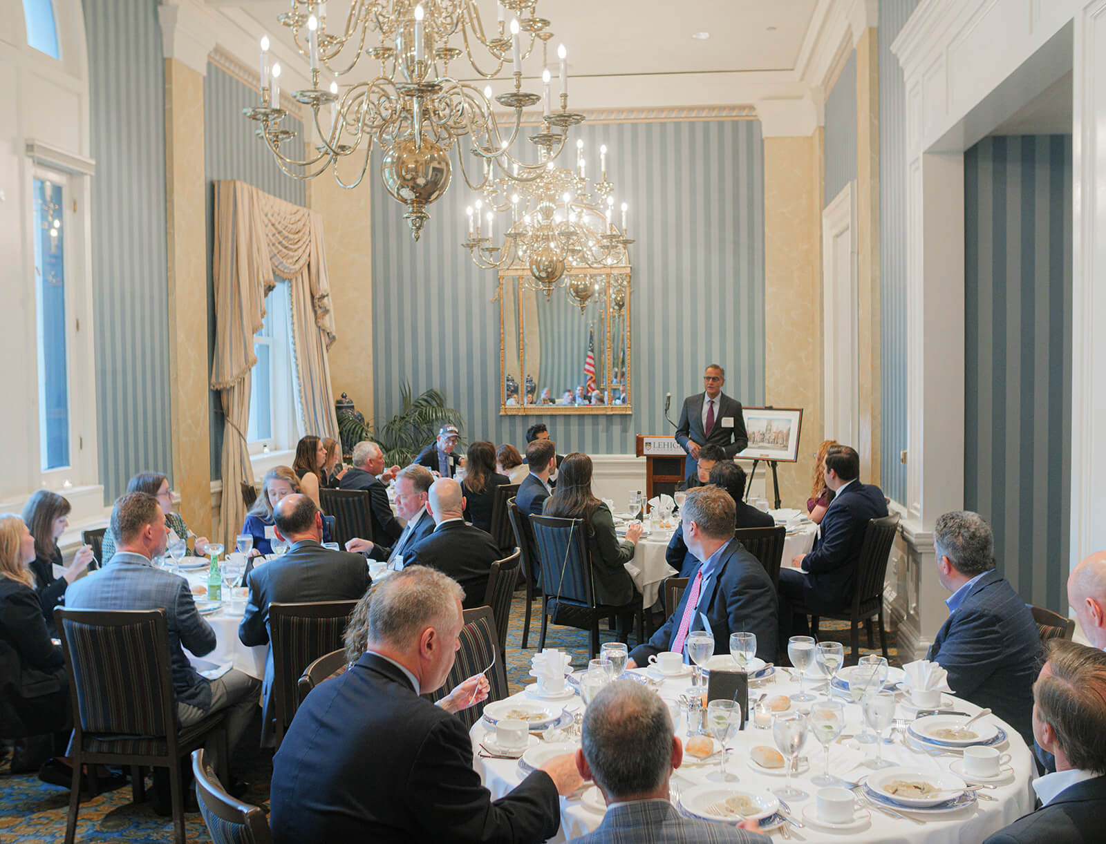 Guests sit in an elegant, well-adorned room with chandeliers and fully set tables while Richard Verma stands in front.