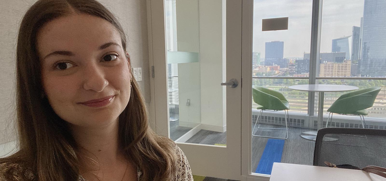 Lauren Lencovich sits at her desk with a view of a skyline behind her