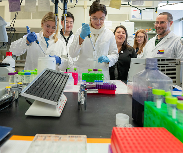 Students in white lab coats and safety glasses use pipettes at a lab bench while others observe and smile in a science lab.