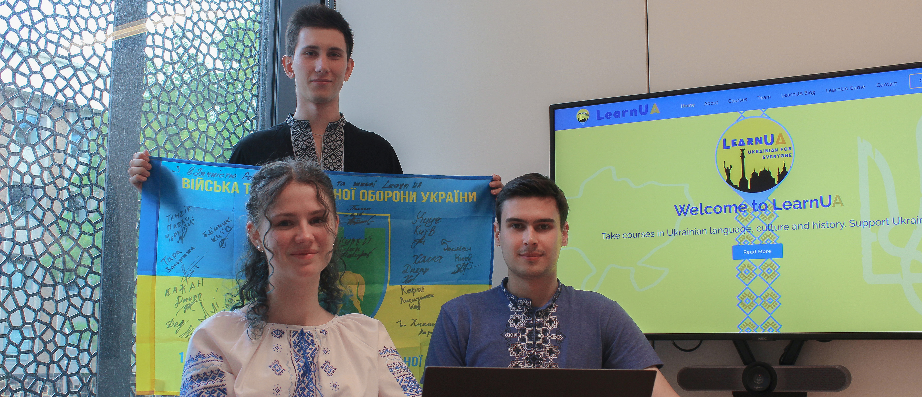 A man and woman sit in front of another man holding a Ukrainian flag while a large screen displays “welcome to LearnUA”