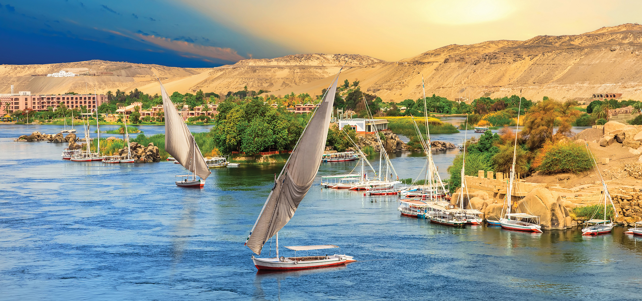 Sail boats float on blue water backed by sandy mountains.