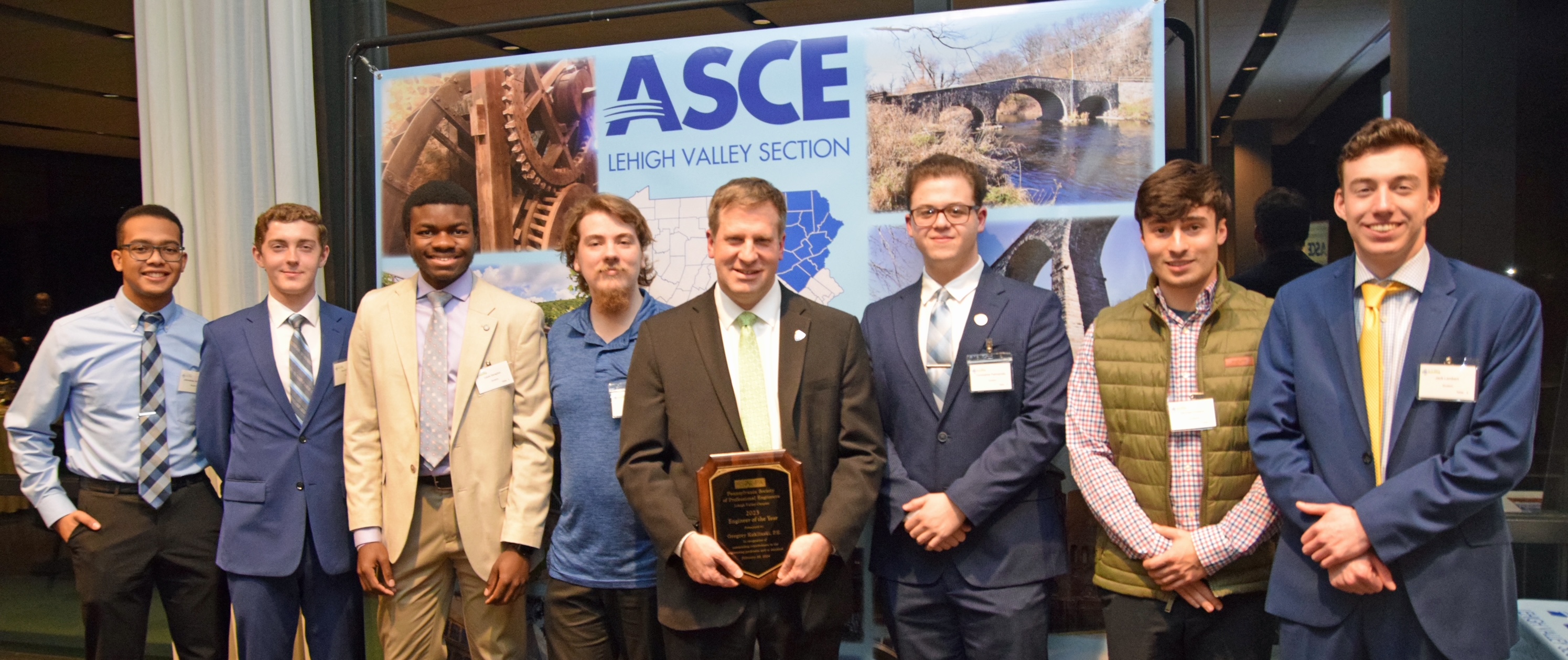 Man holding award plaque stands in from of an "ASCE Lehigh Valley Section" banner flanked by male engineering students wearing business attire and name tags.