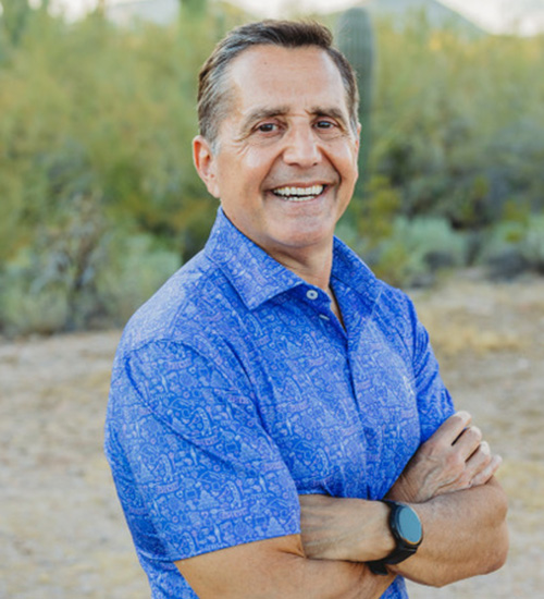 Greg wears a blue polo with desert greenery behind him