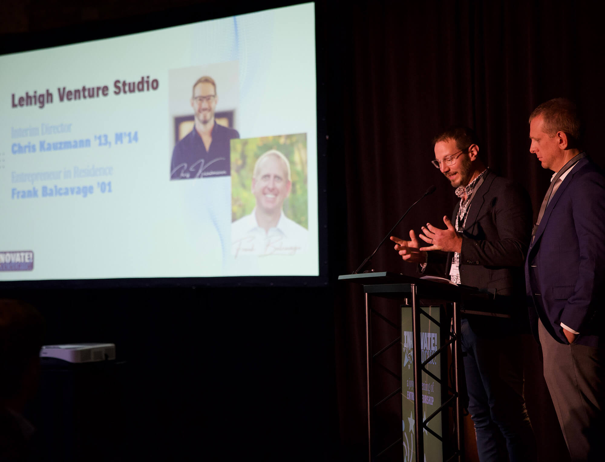 Chris Kauzmann speaks at a podium, motioning with his hands, standing beside Frank Balcavage, with a monitor visible showing both men and their titles under the heading “Lehigh Venture Studio.”