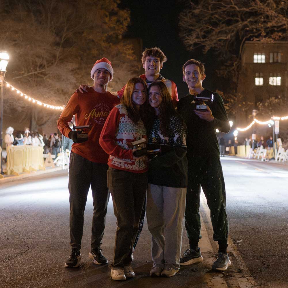 Five young adults pose together outdoors at night on a paved street illuminated by overhead string lights. They are smiling, wearing festive holiday sweaters and pajamas, and holding small awards and trophies, with one person wearing a Santa hat.