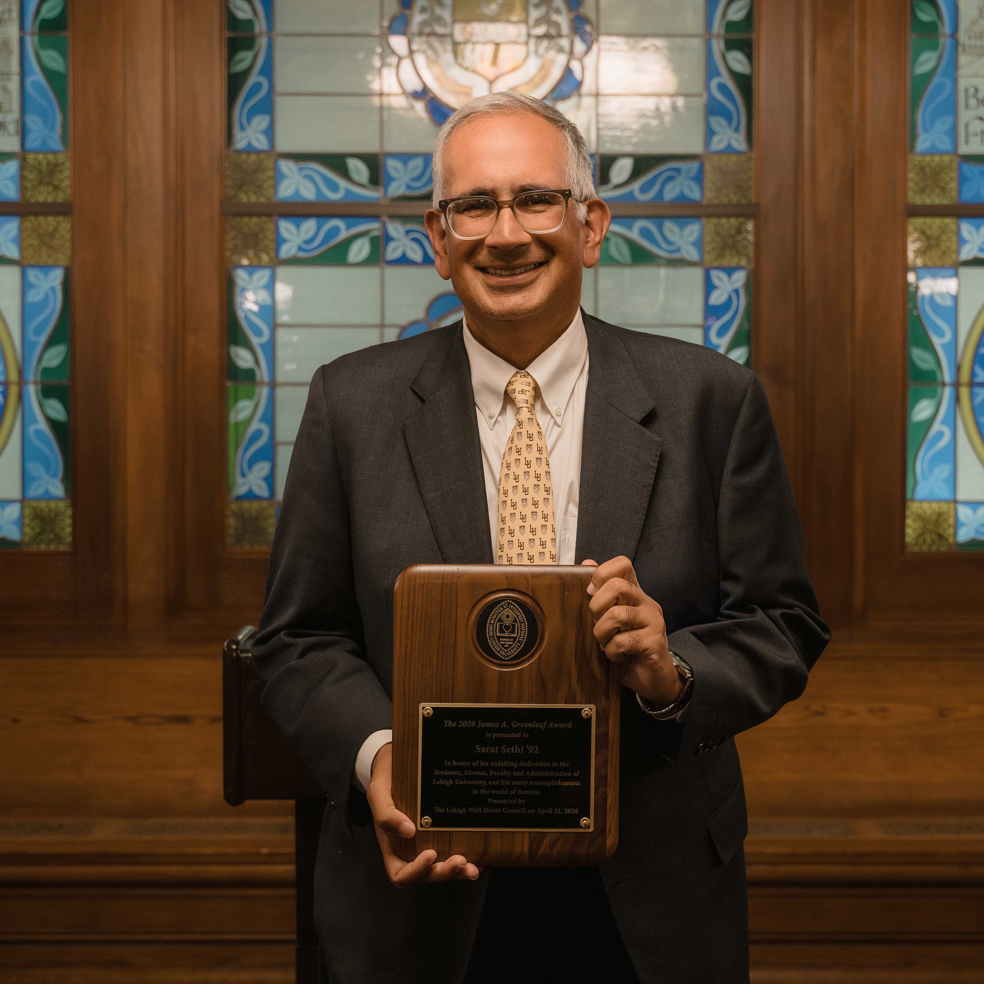 Sarat holds up his award, which is set into a wooden plaque. He wears a gray suit jacket and a yellow tie with Lehigh's logo.