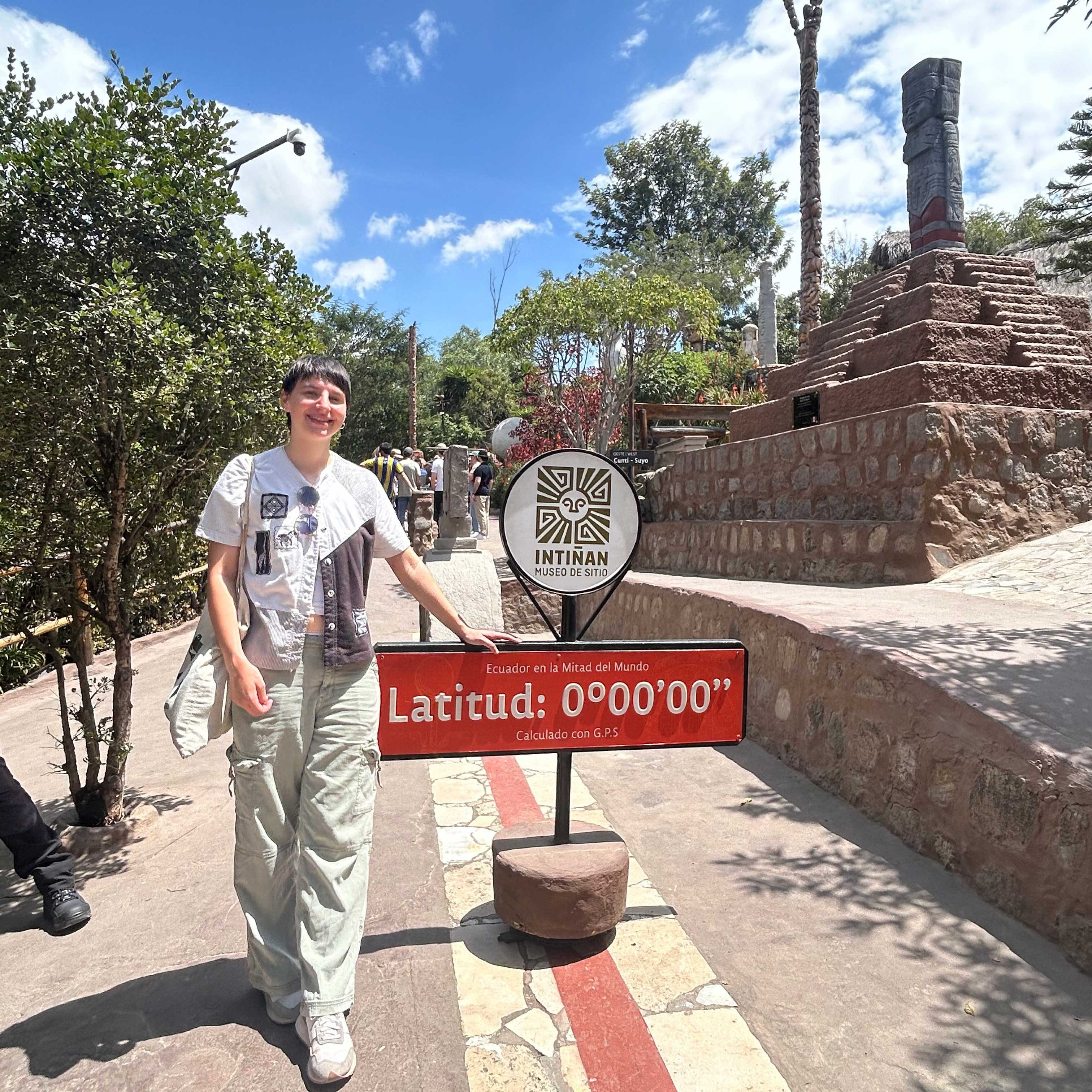 Lillian stands alongside a sign marking the equator while wearing light cargos and a white top.