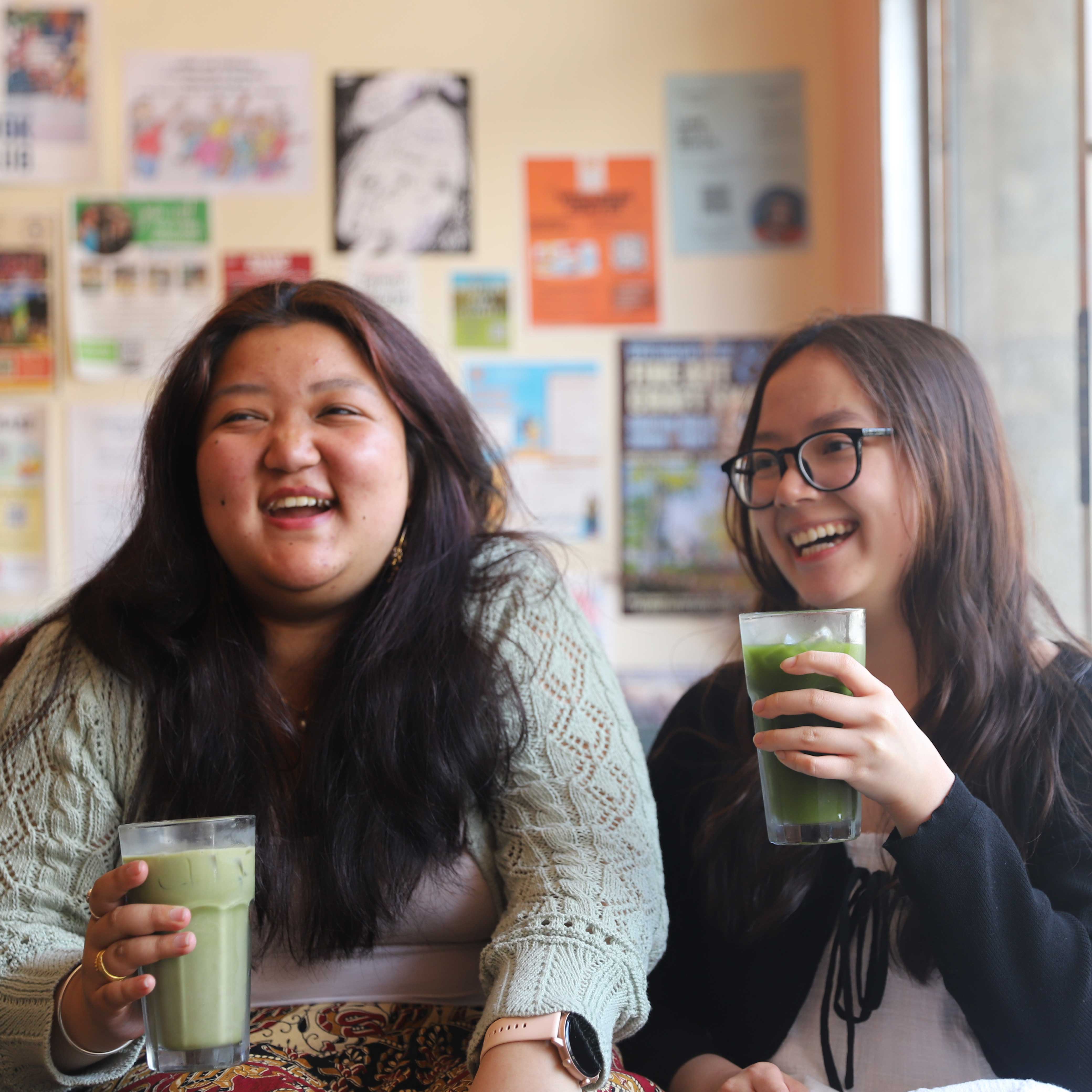 Two young women laugh while holding iced matcha lattes in their hands.