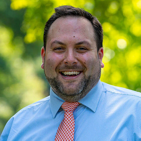 A headshot of Scott Wojciechowski, standing outside against a blurred green landscape and wearing a blue shirt with orange tie.