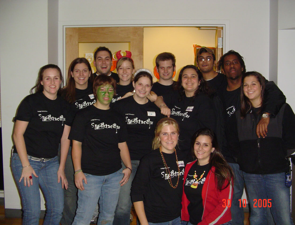 Group of college students wearing matching black Spooktacular shirts pose for a photo, one wearing green face paint.