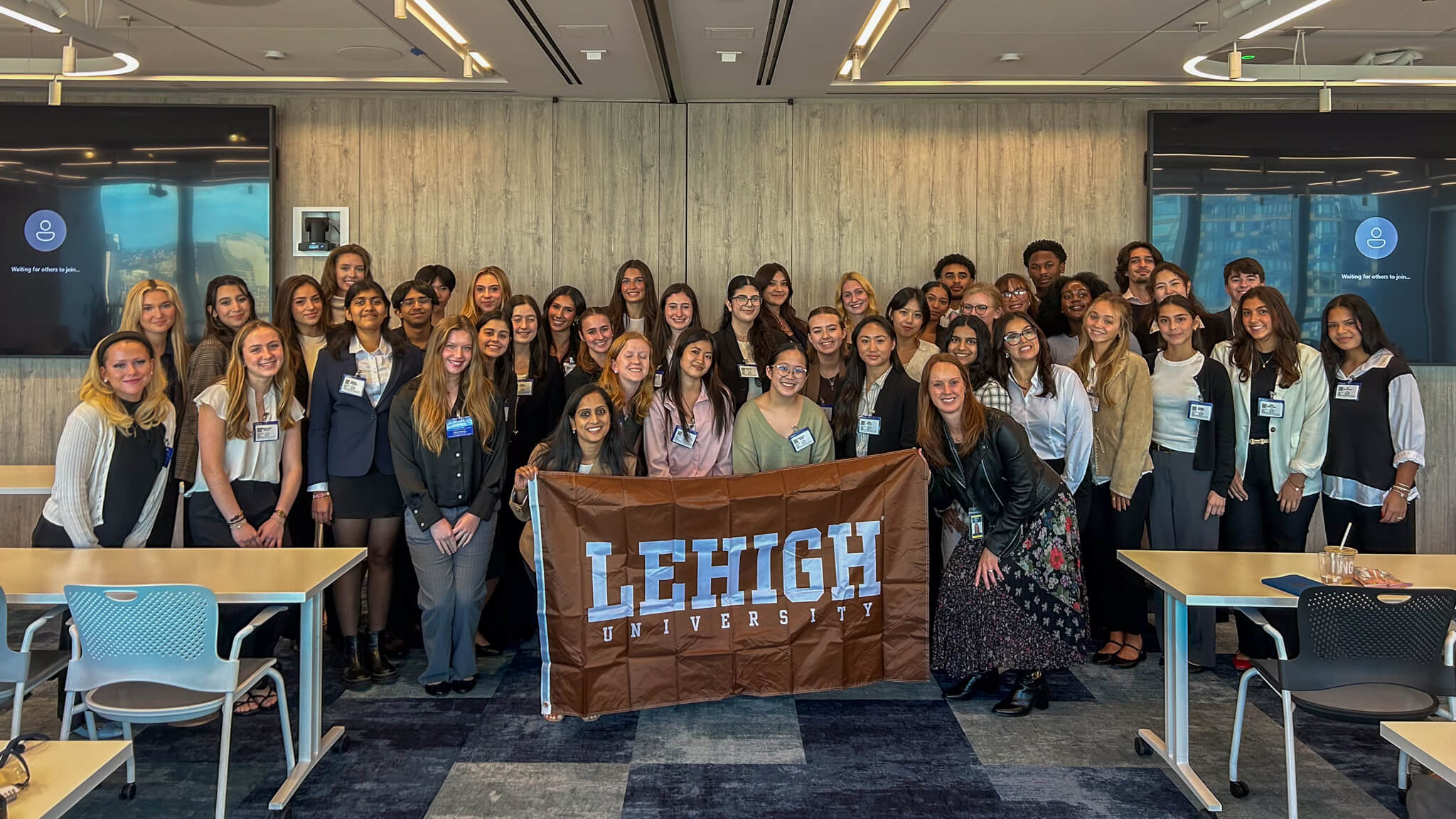 A large group of students stand together and smile. Two Lehigh alumni near the front hold up a brown Lehigh banner.