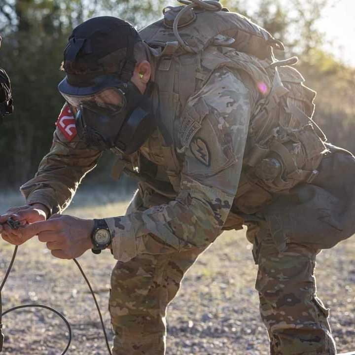 Lucas wears his military fatigues, backpack, and a black gas mask as he bends and works on a set of wires. 