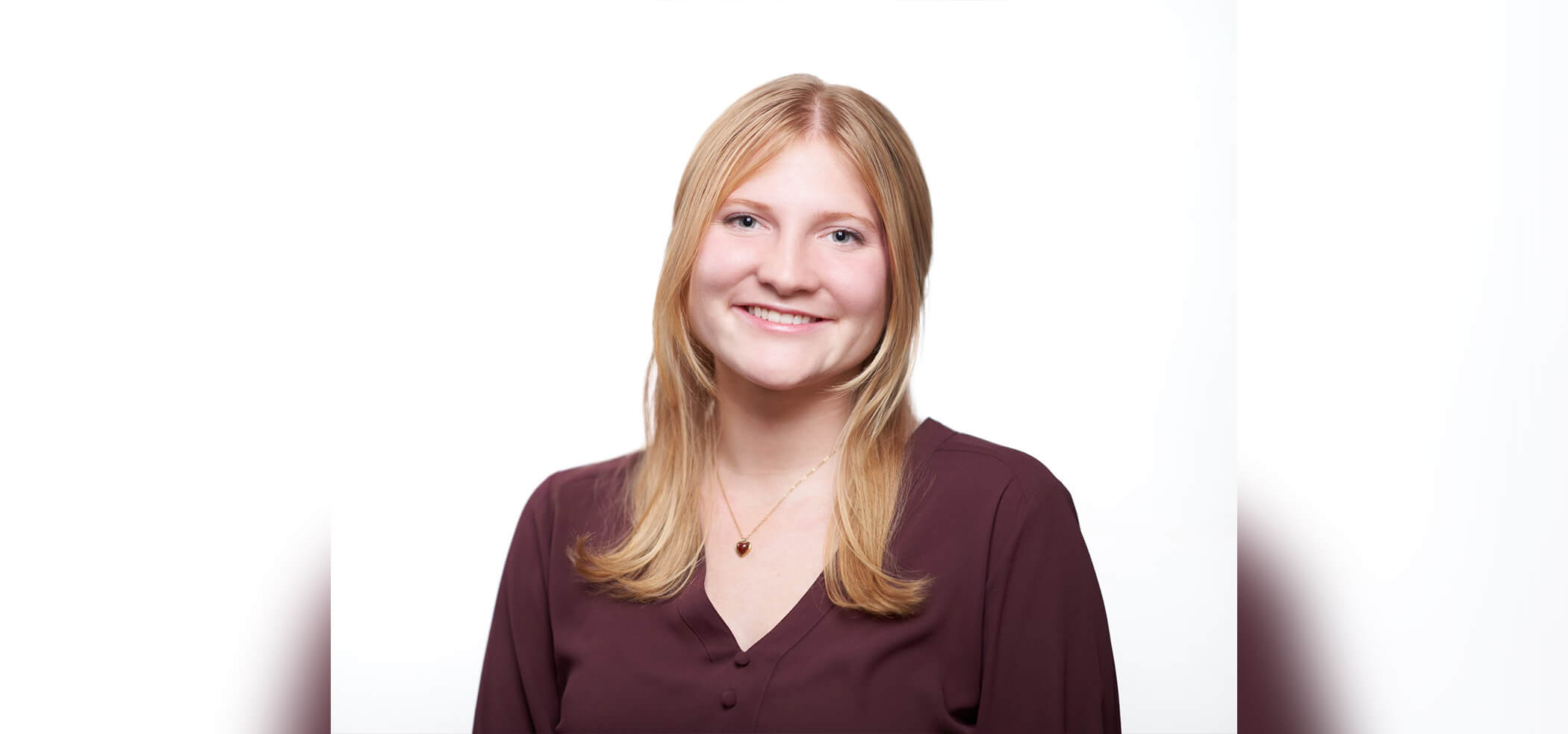 A woman smiles against a white backdrop wearing a maroon v-neck shirt and a small pendant with strawberry blonde hair worn down.