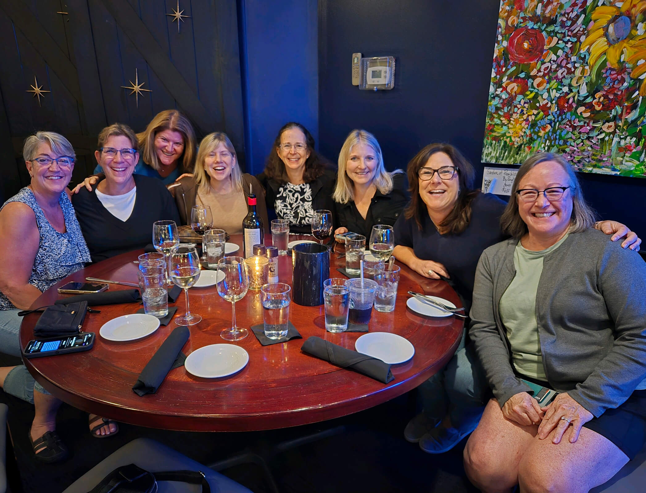 8 women huddle around a round table in a dimly lit restaurant, sliming and laughing as they pose for a picture.