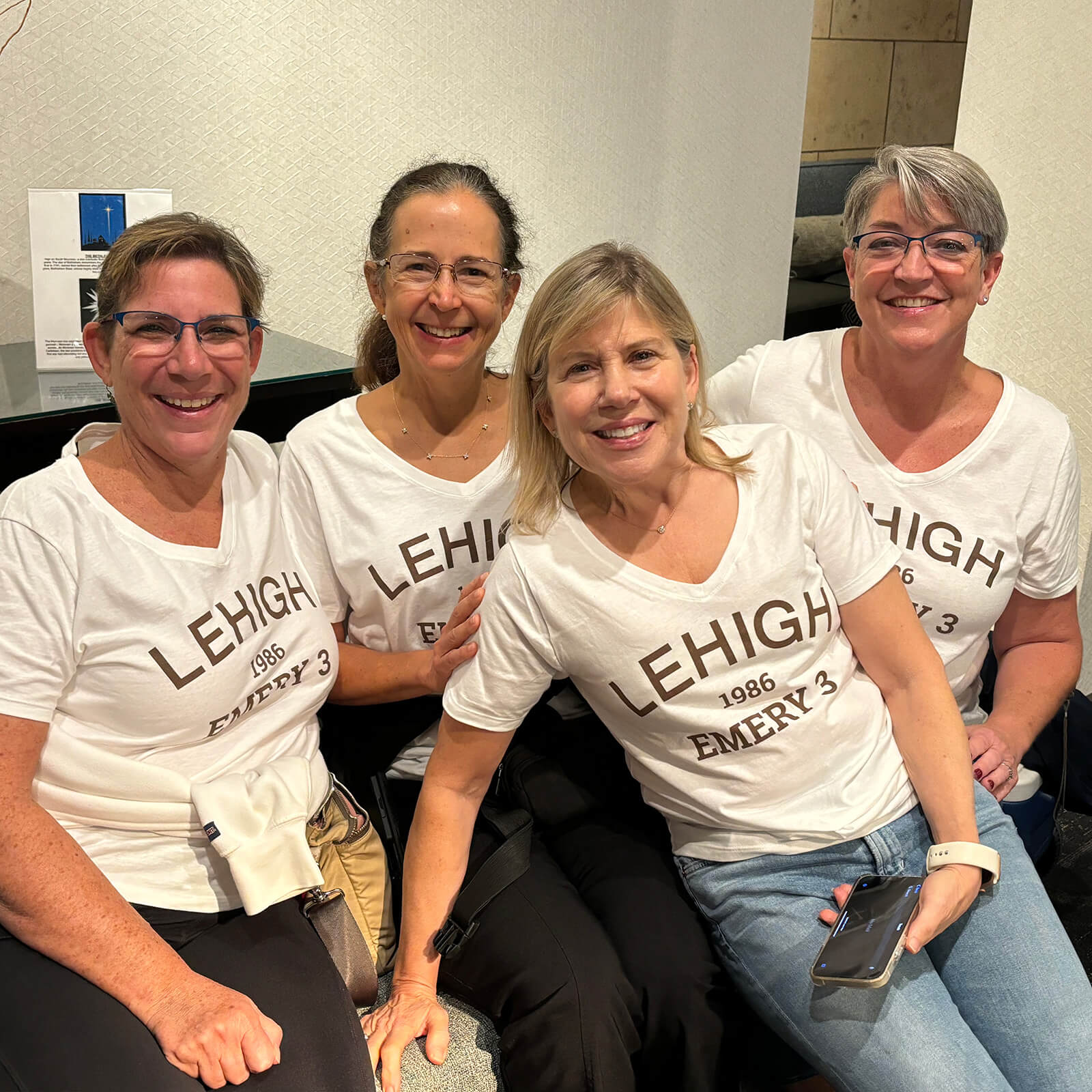 4 women sit closely together smiling for a photograph and all wear the same white tshirt that says "Lehigh 1986 Emery 3".