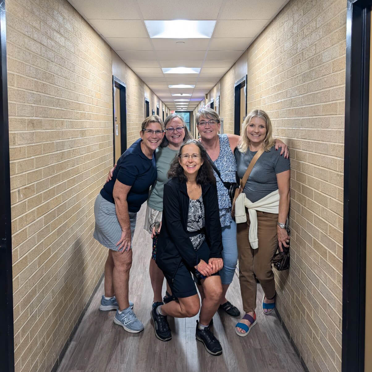 Five women pose in the hallway of Emery, with brick walls lining the corridor and drop ceiling tiles and fluorescent lights above them.