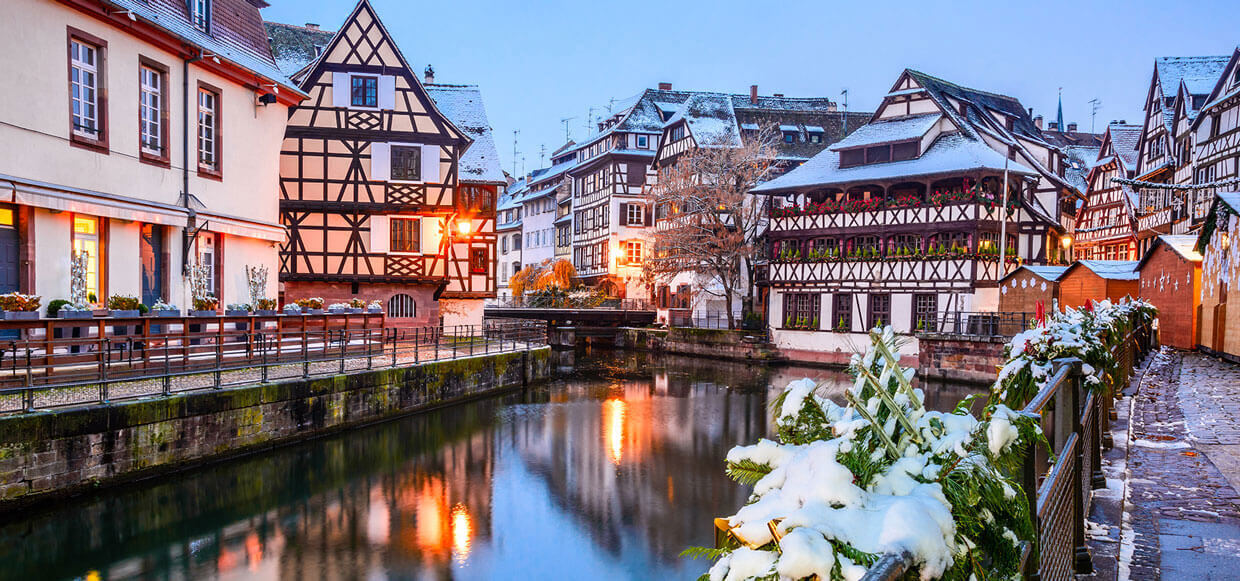 Snow-covered walkways and buildings lining a river at dusk.