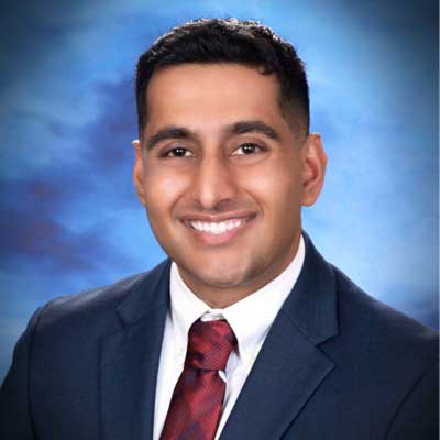 Maanik wears a navy suit jacket with a white button up and a red tie. He stands in front of a blue background and smiles.