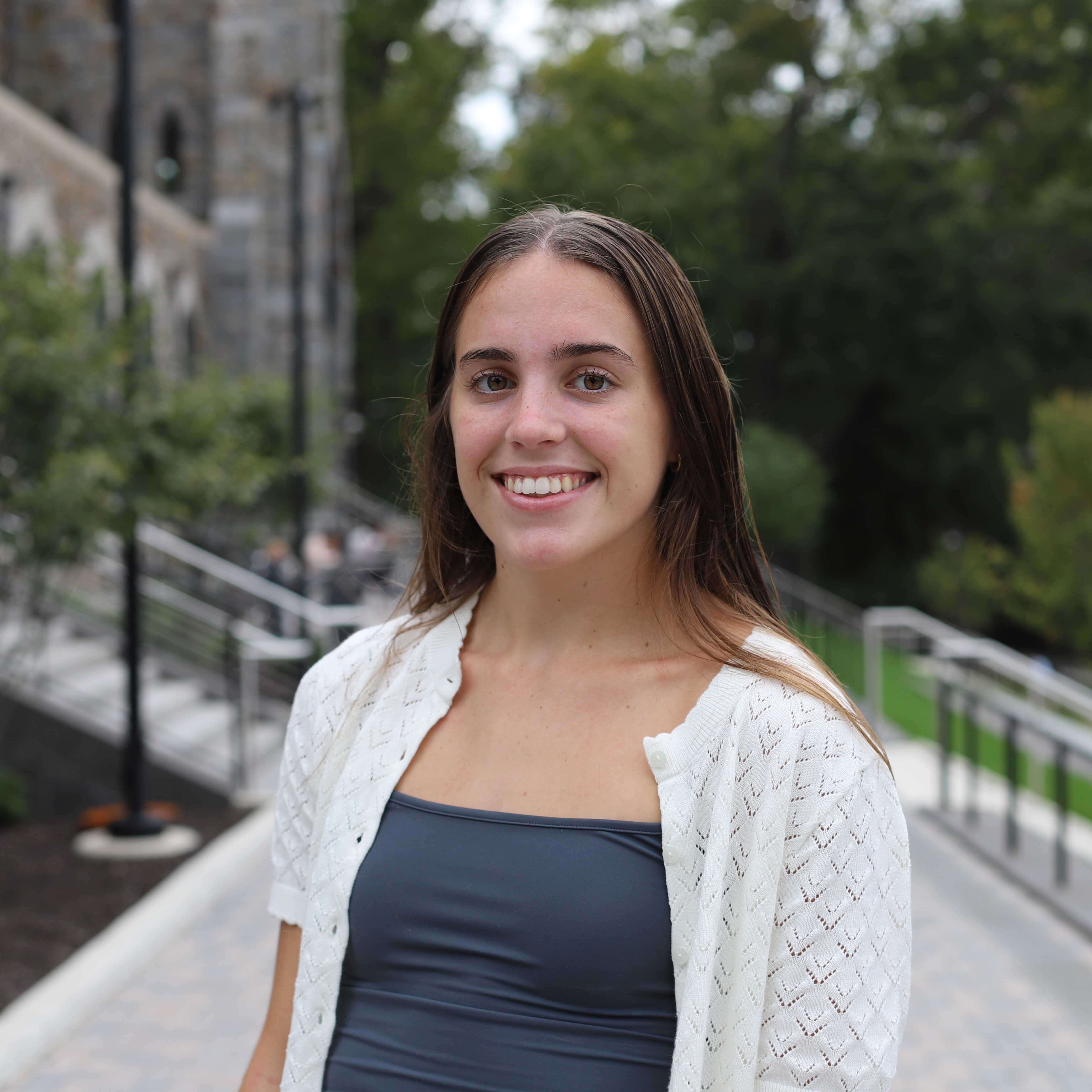 Maite wears a grey camisole with a cream, short-sleeved cardigan atop. The Clayton University Center steps are visible behind her.