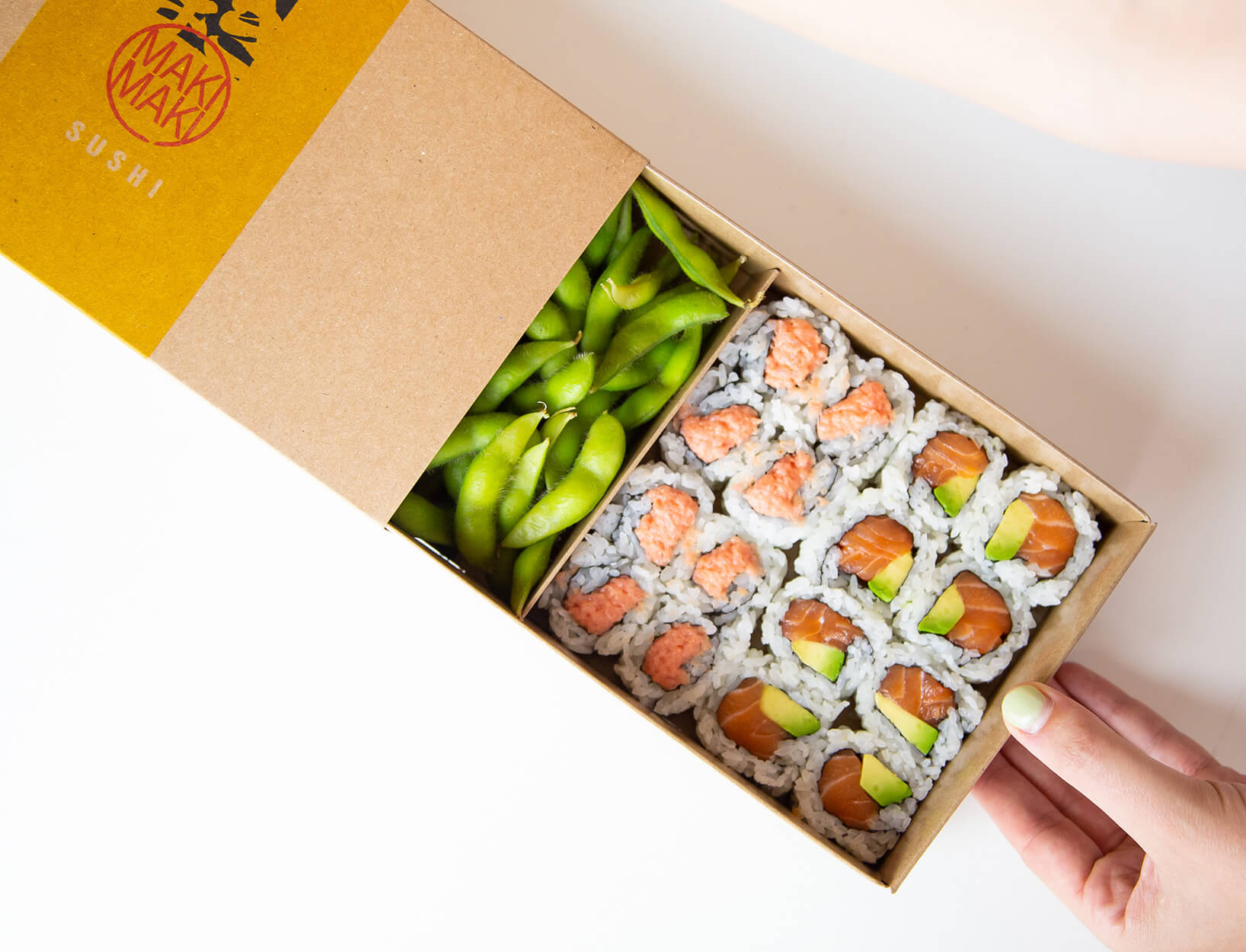 A modernly designed brown sushi takeout box is filled with two types of rolls and edamame.