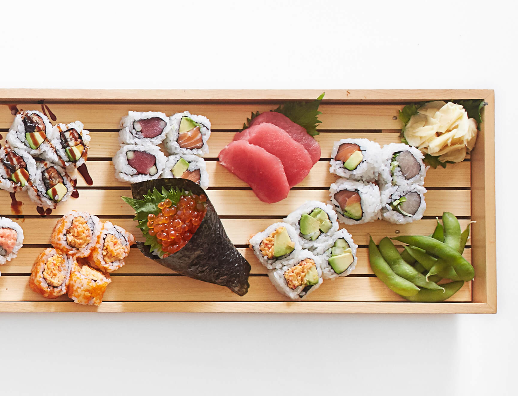 A bamboo tray features an assortment of traditional sushi and rolls, garnished with ginger and edamame.