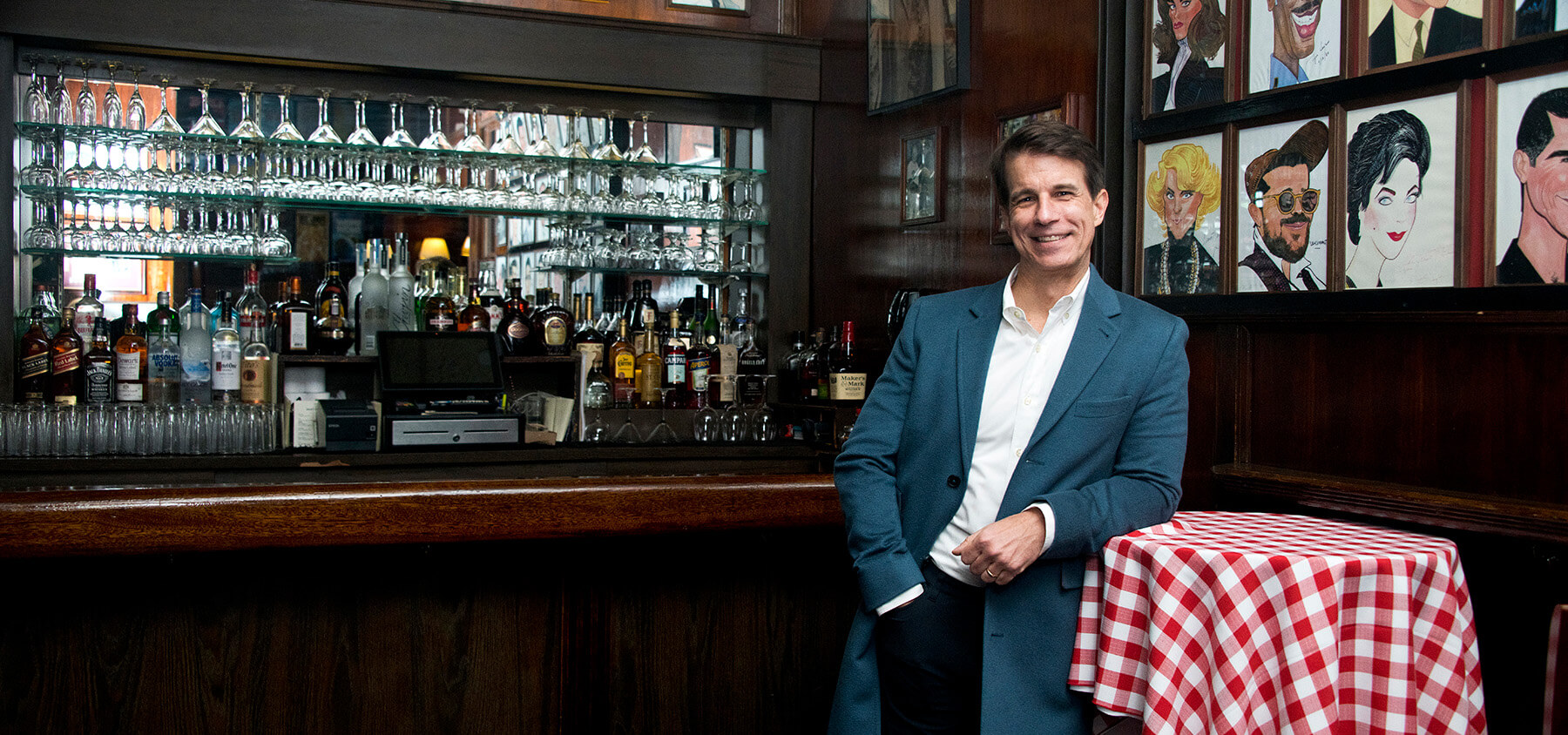 Wearing a suit jacket, a man smiles beside a bar, leaning on a hightop table draped with a red and white checkered table cloth.