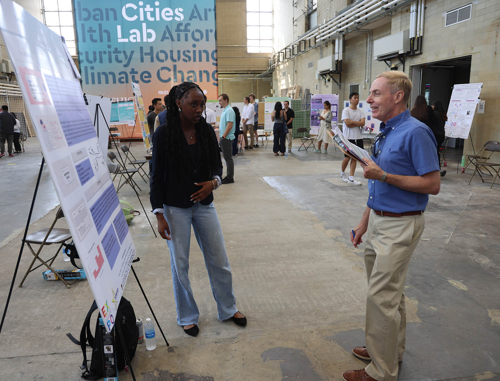 Mark Shiner holding a clipboard stands in front of a student and her presentation board as she explains her research.