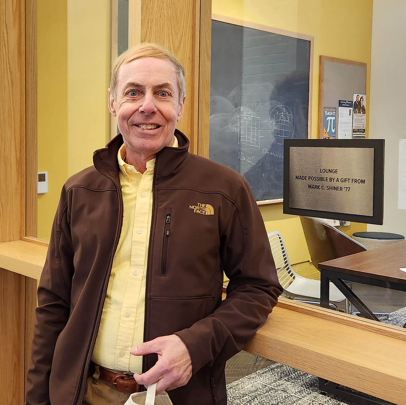 Mark Shiner stands beside a window to a lounge with a plaque bearing his name and class year, recognizing his charitable gift.