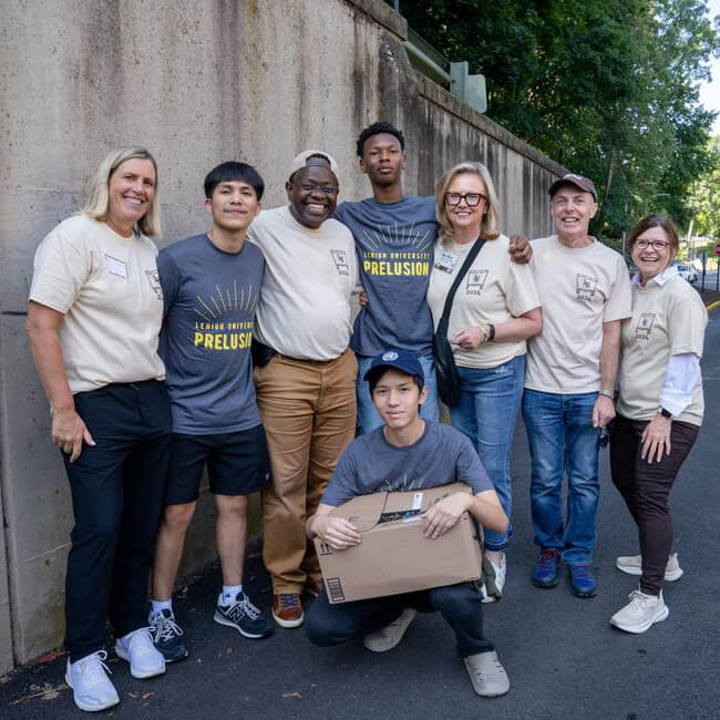 A group of alumni stand with students and faculty in front of a concrete wall, volunteering to help with move in.