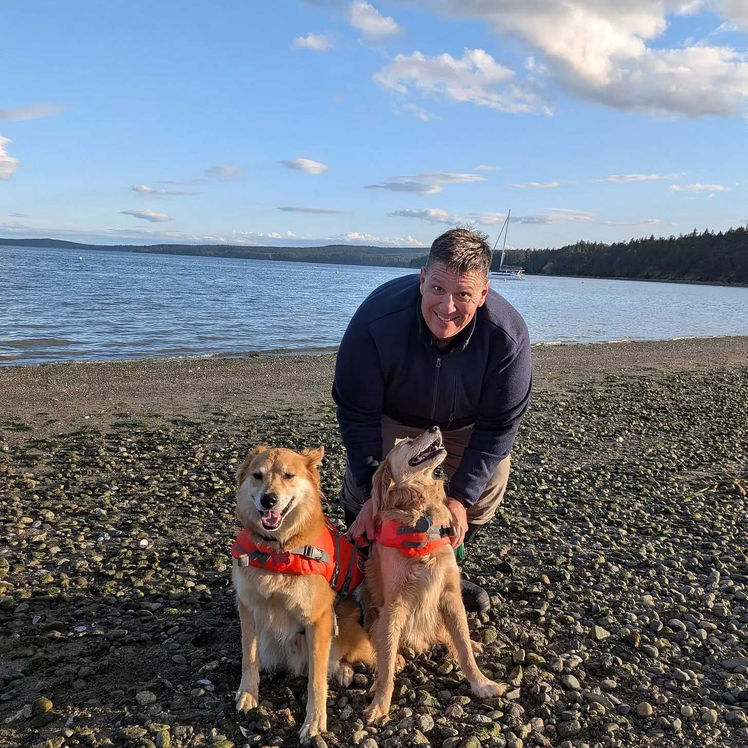 Mark bends down and pets two dogs on a pebble beach with clear water and trees visible behind him.