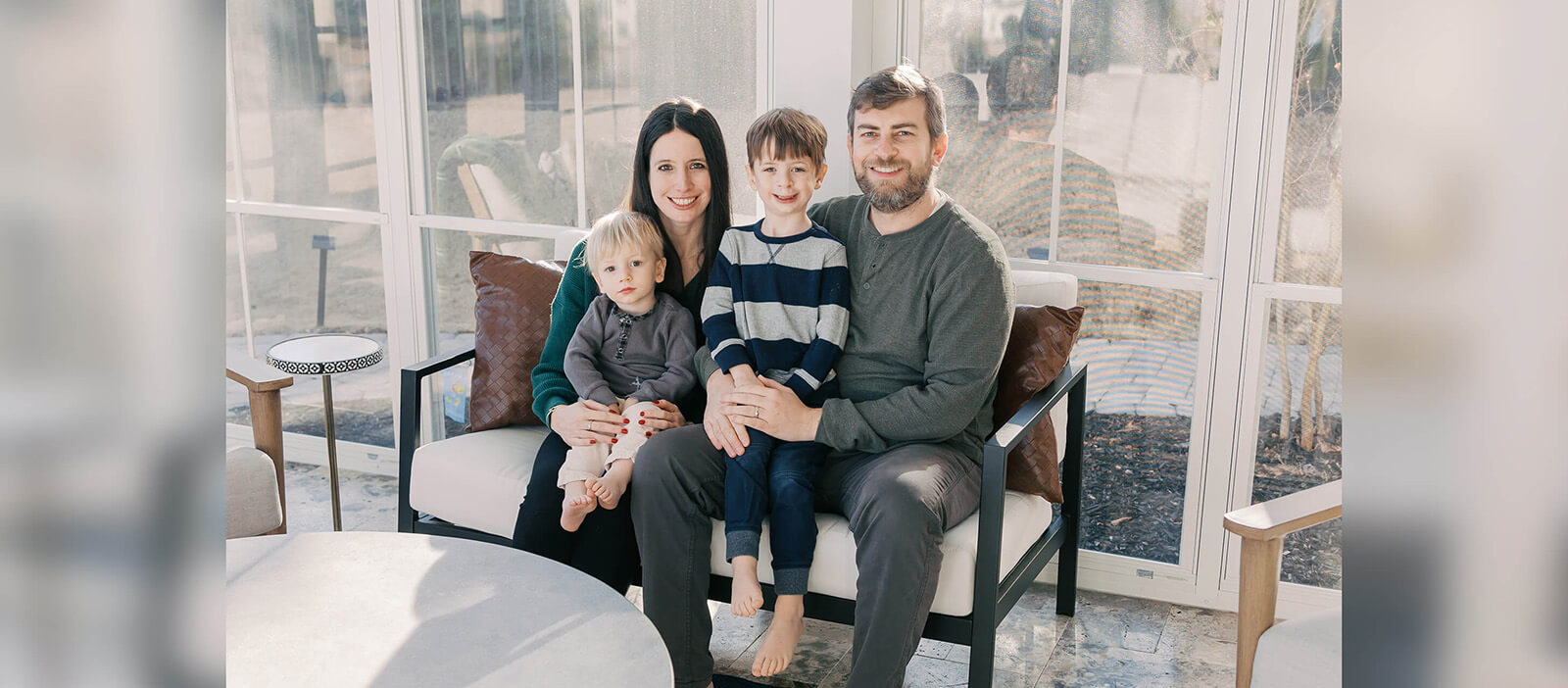 Family sits for a portrait in a well lit white room against a wall of windows, with father and mother each holding a small boy in their lap.