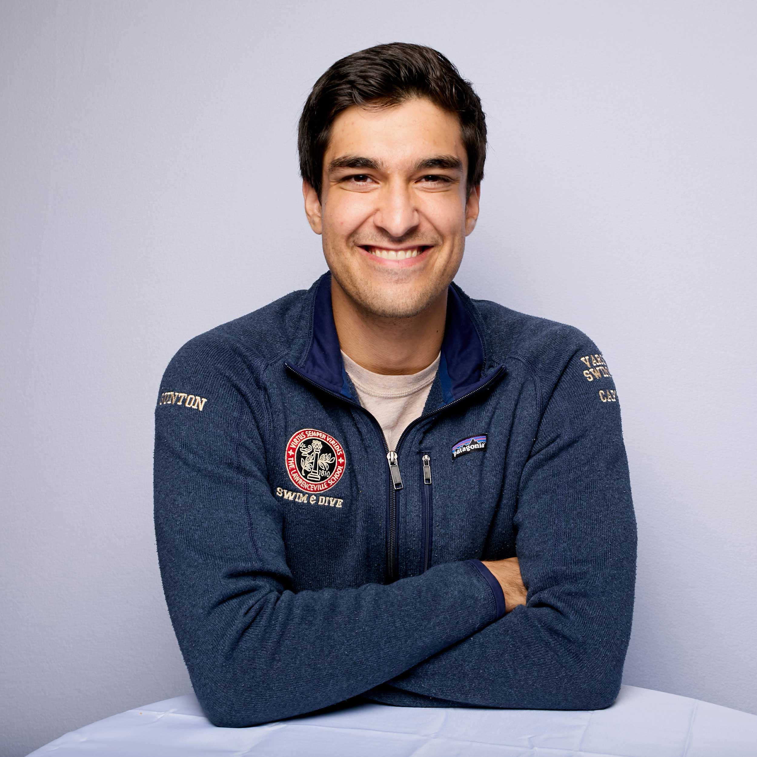 Matthew wears a navy zip-up sweater with a beige shirt visible underneath. He smiles and sits at table that his elbows rest on.