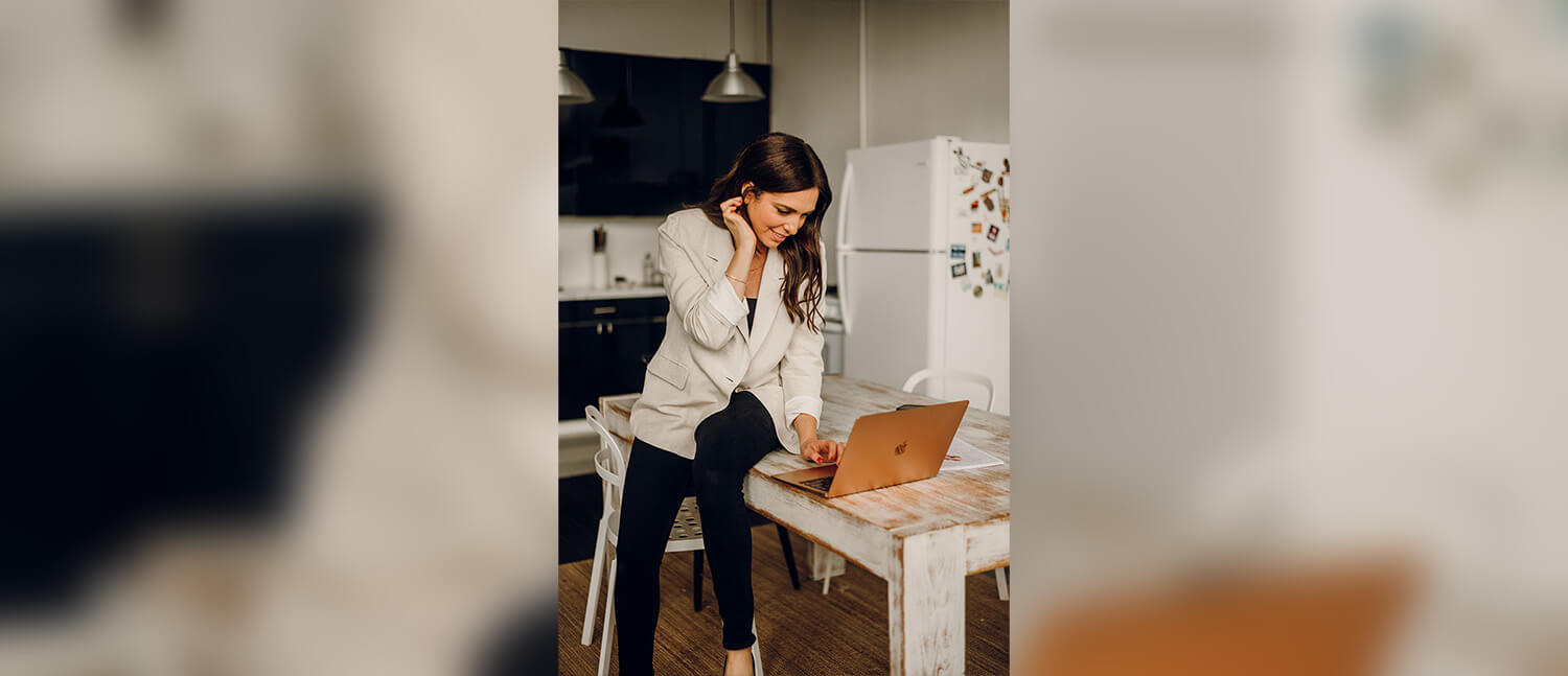 Maura Walters sits on a table in a kitchen and uses a laptop while talking on the phone.