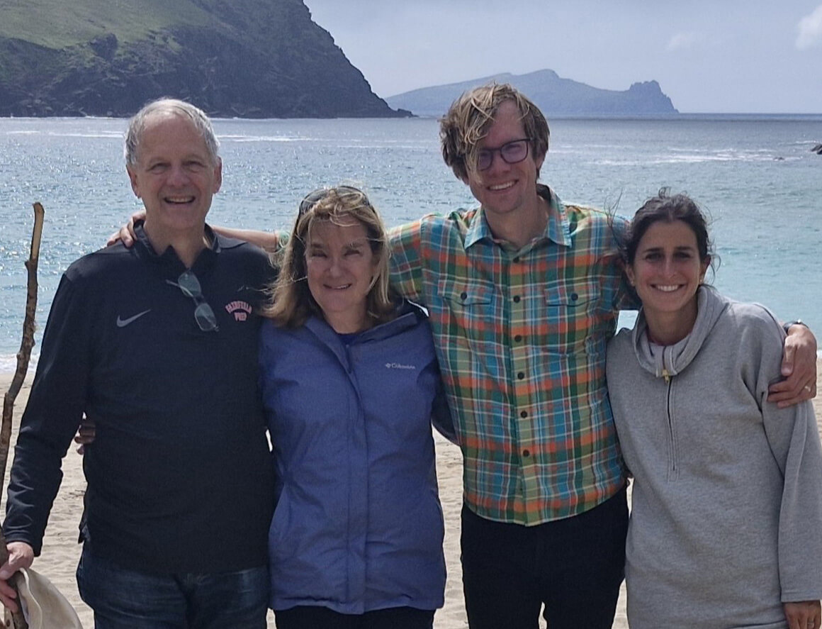 McLeod family poses smiling shoulder to shoulder, arm in arm, on a beach in front of water with mountains in the distance.