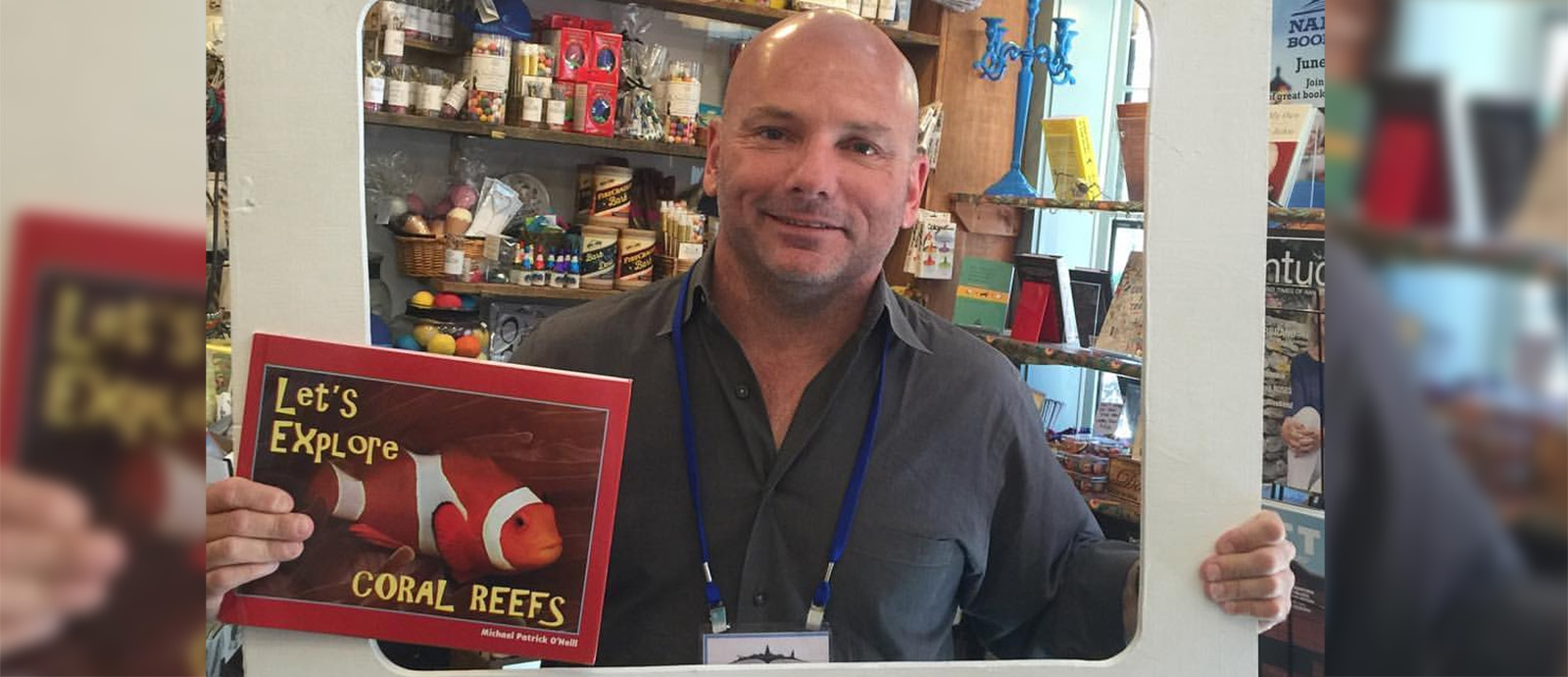 Michael Patrick O’Neill standing in a gift shop holding his book titled Coral Reefs.