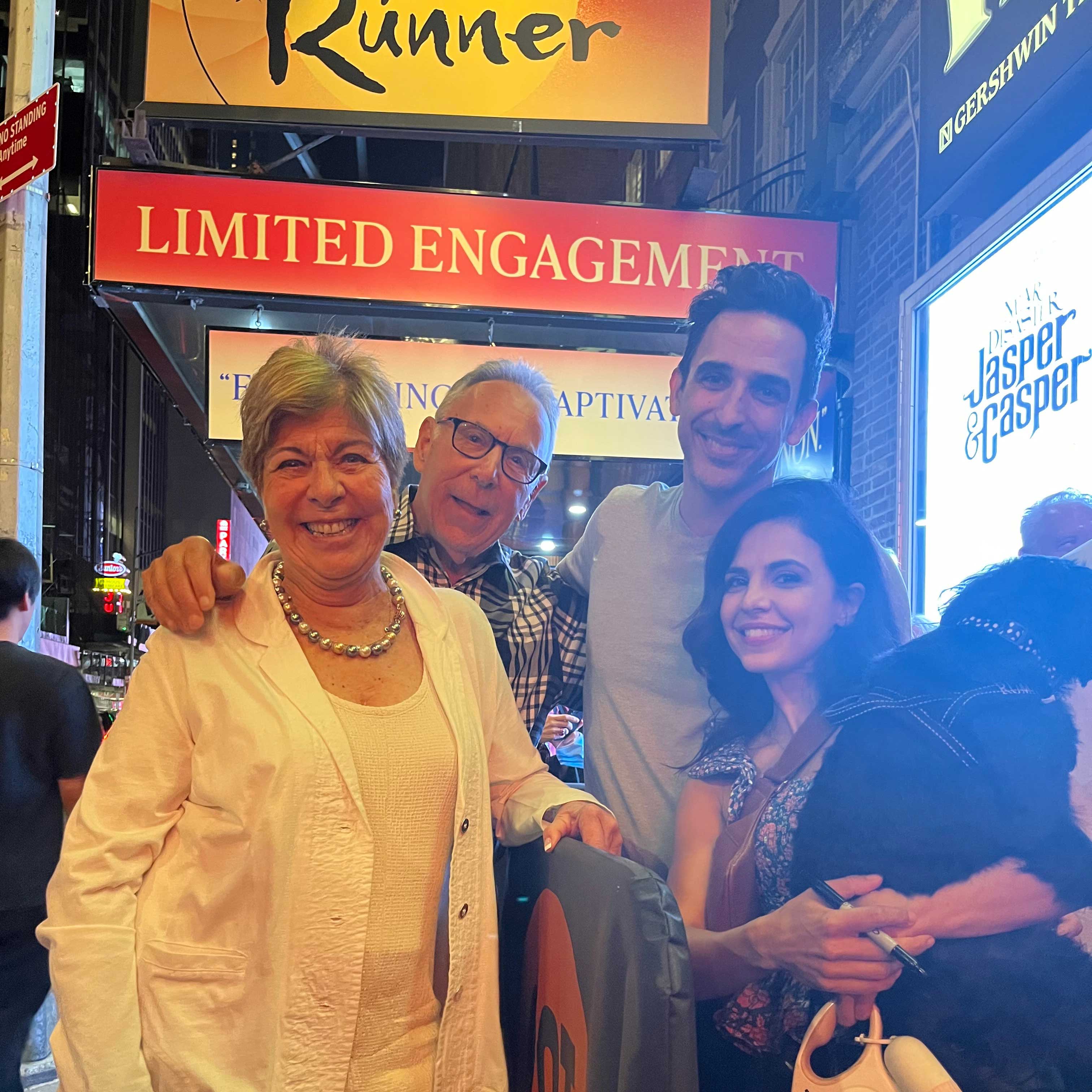 Next to a man and a woman who is holding a dog in her arms, Michael and Enid Seiden stand outside of the theater with The Kite Runner marquee hanging above them.