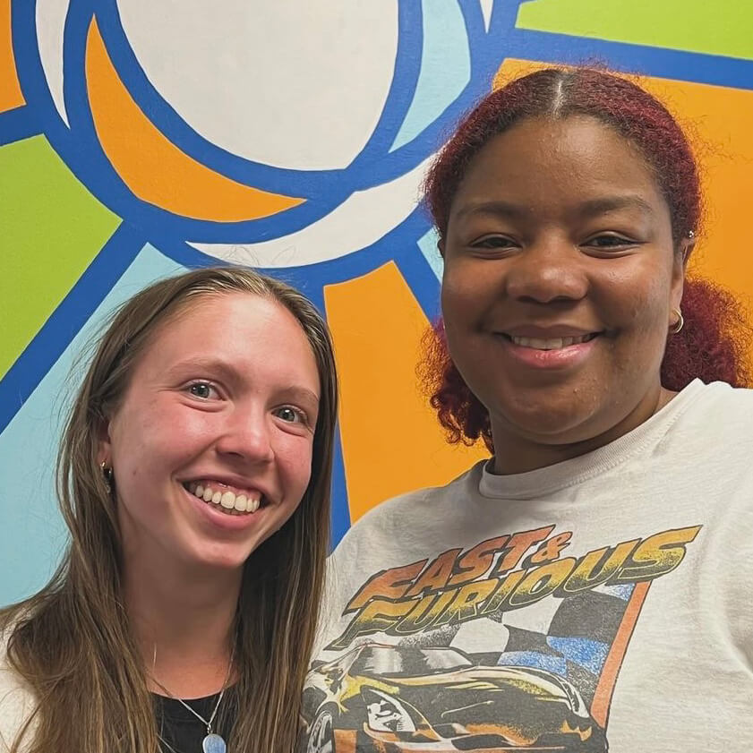 Two female students stand smiling in front of a brightly colored mural.