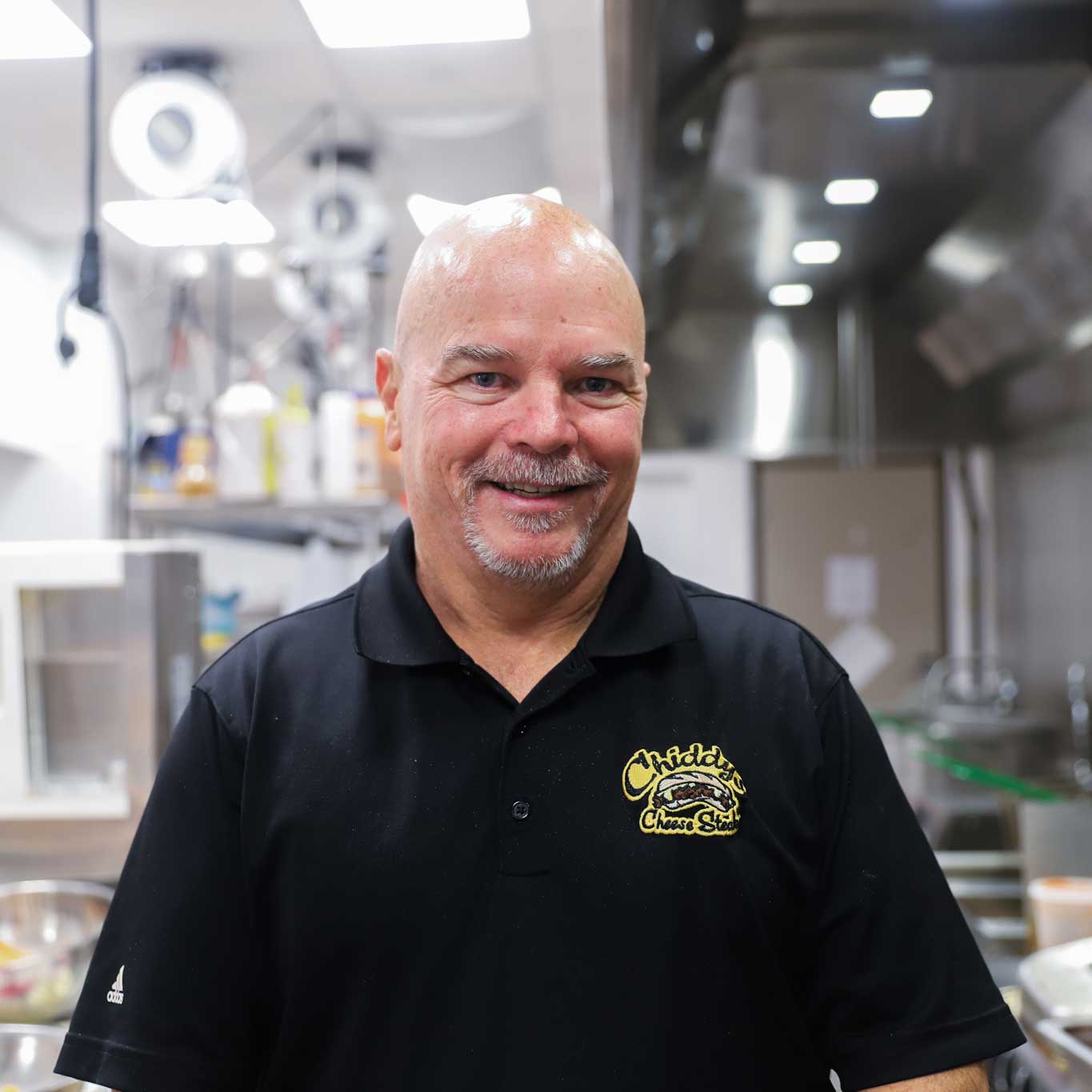 Michael Chidester wears a black, branded Chiddy's Cheesesteak's shirt and smiles while standing in an industrial kitchen.