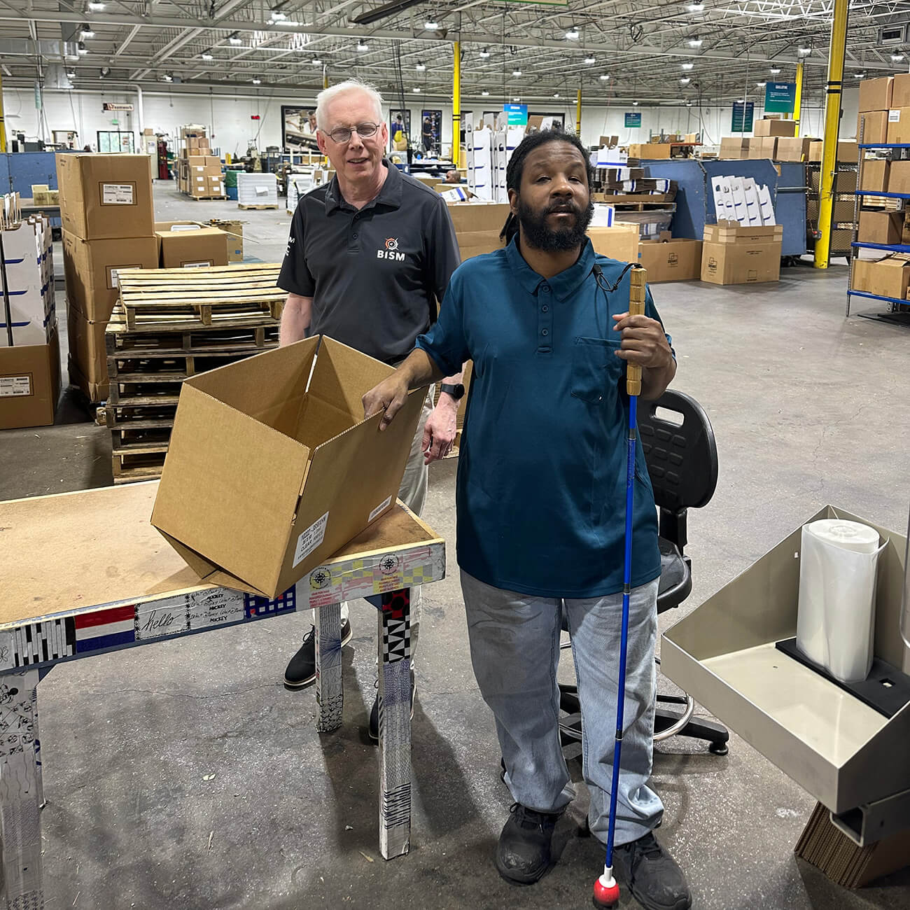 Mike Gosse stands at a table in a warehouse alongside a man holding a brown box in one hand and a cane in the other.