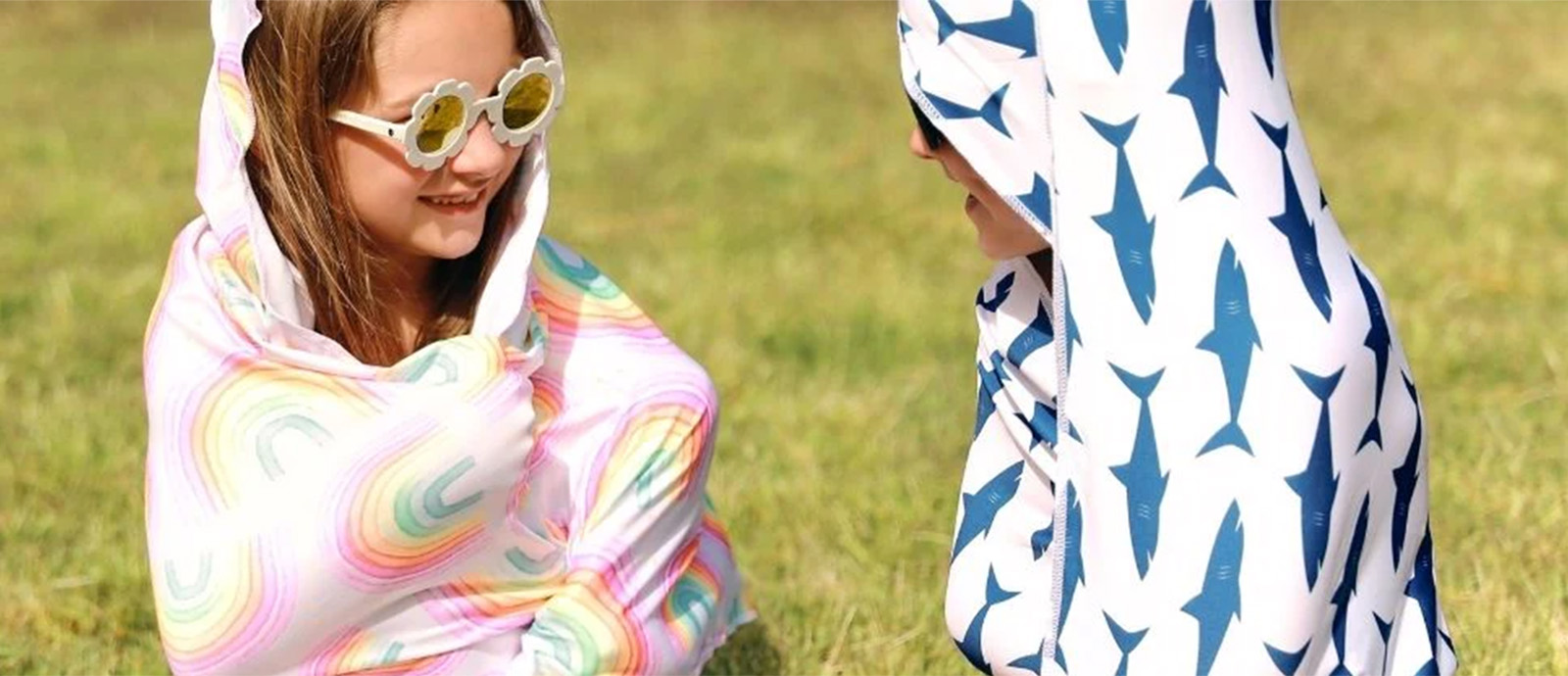 A young girl and boy in sunglasses wrapped in rainbow and shark print towels.